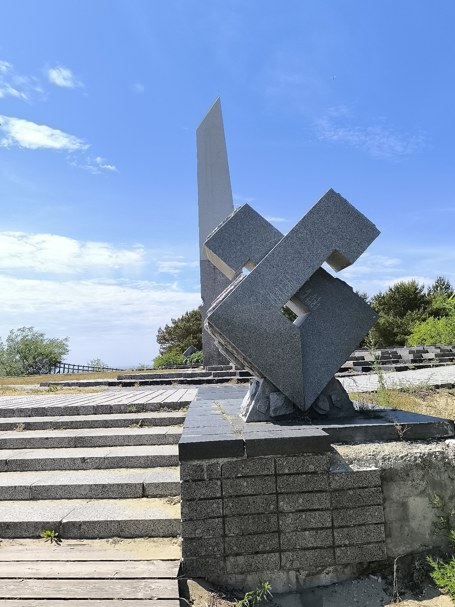 The image shows a gray granite monument with abstract geometric shapes, located outdoors on a sunny day. The monument is situated on a stone staircase with a wooden walkway in front. In the background, there are trees, a body of water in the distance, and a blue sky with scattered white clouds. There are no people visible in the image. This monument is located in Neringa, Lithuania.