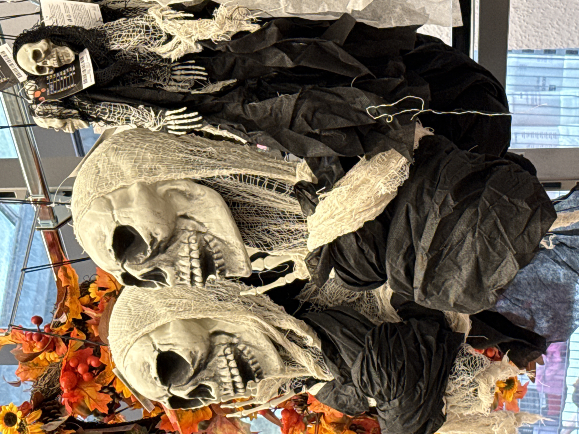 The image displays a close-up view of Halloween decorations, specifically several skeletal figures hanging in what appears to be a store or market setting. The figures are designed to resemble grim reapers or ghosts, with prominent skull heads, draped in tattered black and white cloth. One skull on the left is partially obscured by a leafy fall garland featuring red berries and sunflowers. The backdrop suggests an indoor environment with metal shelving and possibly windows with blinds. The overall color palette is dominated by the muted tones of the decorations, contrasted with the bright fall foliage and the blur of the background. The specific location in Bad Lauterberg im Harz, Germany, is not evident from the visual information provided in the image.