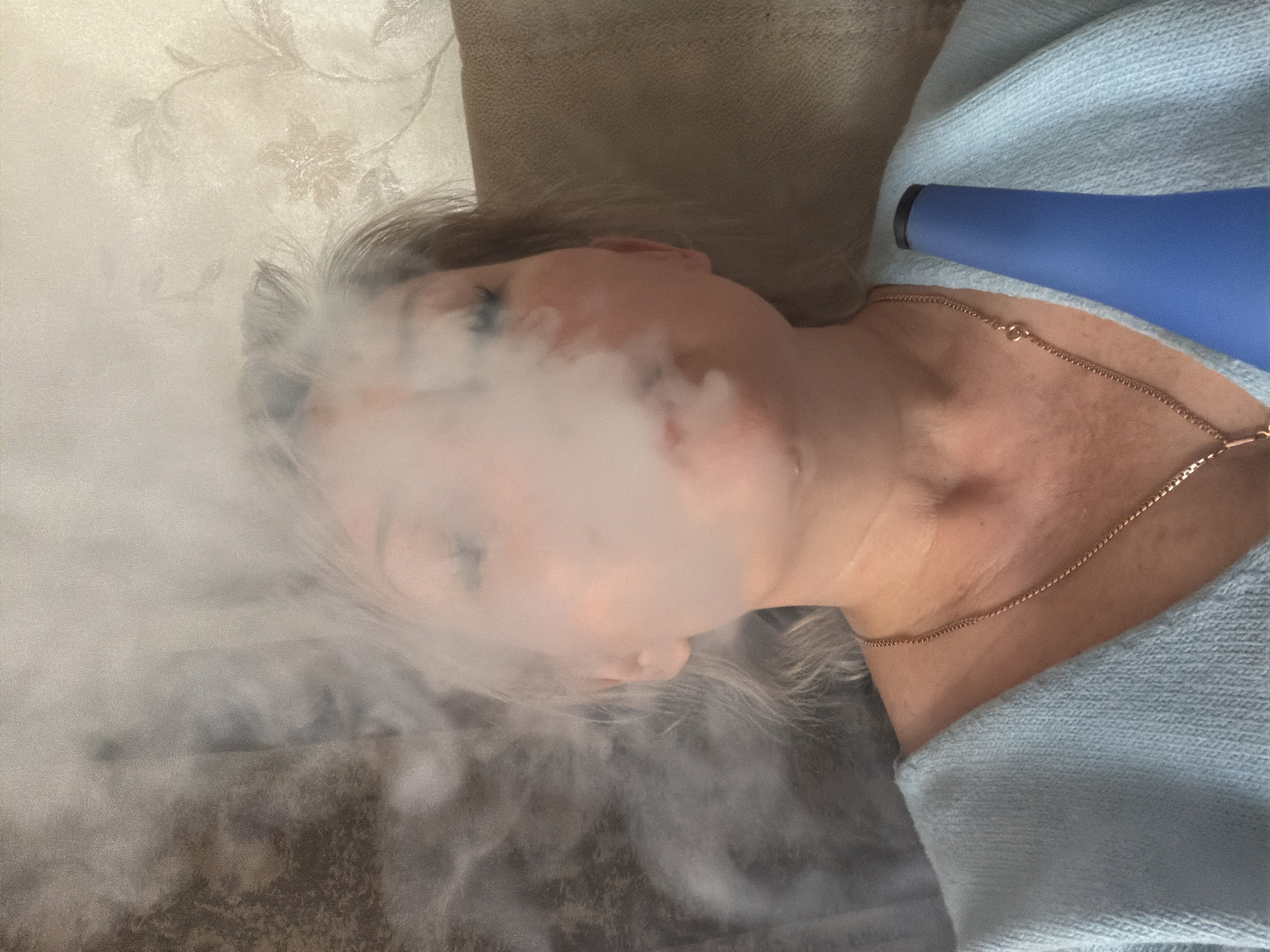 A Caucasian female reclines, enveloped in white smoke. She wears a light blue garment and a gold necklace. A blue object, possibly a device, is positioned near her neck. The background includes a beige couch and a patterned wallpaper. The location is unknown.
