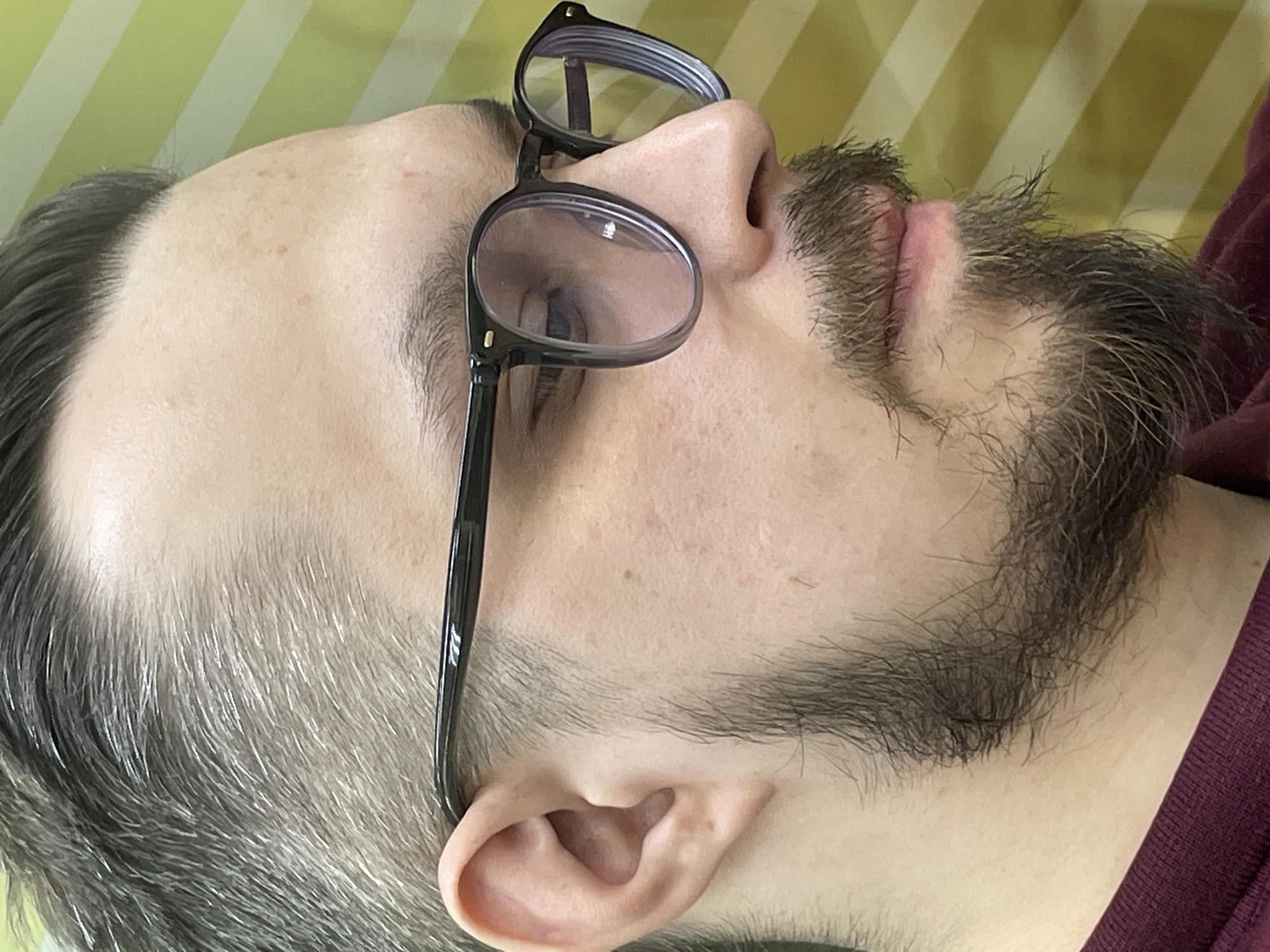 A close-up, profile shot taken in Krasnoyarsk, Russia, features a man lying down indoors. He has fair skin, a short beard, and short hair that transitions from a darker tone at the front to a prominent mix of gray and silver strands on the sides, extending into his beard and sideburns. He wears dark-framed eyeglasses, perched on his nose, with his left eye partially visible through the lens, looking upwards or to the side. His expression is neutral. His lips are slightly parted, showing a reddish hue.
The background consists of an alternating striped pattern in light green/yellow and white/cream, suggestive of bedding or a pillow, indicating an indoor setting. A burgundy or maroon garment is visible around his neck and shoulder. Small brown spots or freckles are present on his forehead. The man appears to be resting or in a contemplative state. No visible text is present in the image.