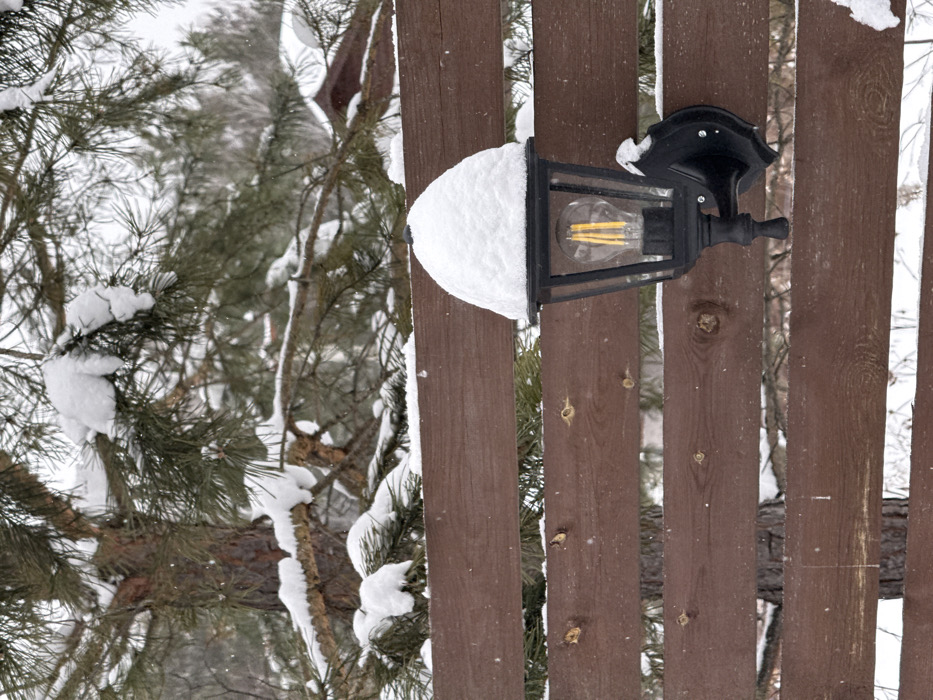 This image, captured in Sysert, Russia, presents a winter outdoor scene. A dark brown wooden fence, constructed from horizontal planks, traverses the midground. Attached to the fence is a black, wall-mounted outdoor lantern. The lantern's upper surface is entirely covered by a thick, uniform layer of white snow. Inside the lantern, a single clear LED filament bulb is visible, emitting a warm, yellowish light. The background features several snow-dusted green pine trees, partially obscured by the fence. Subtle white flecks, indicative of ongoing snowfall, are dispersed throughout the frame. No persons or overt activity are depicted.
