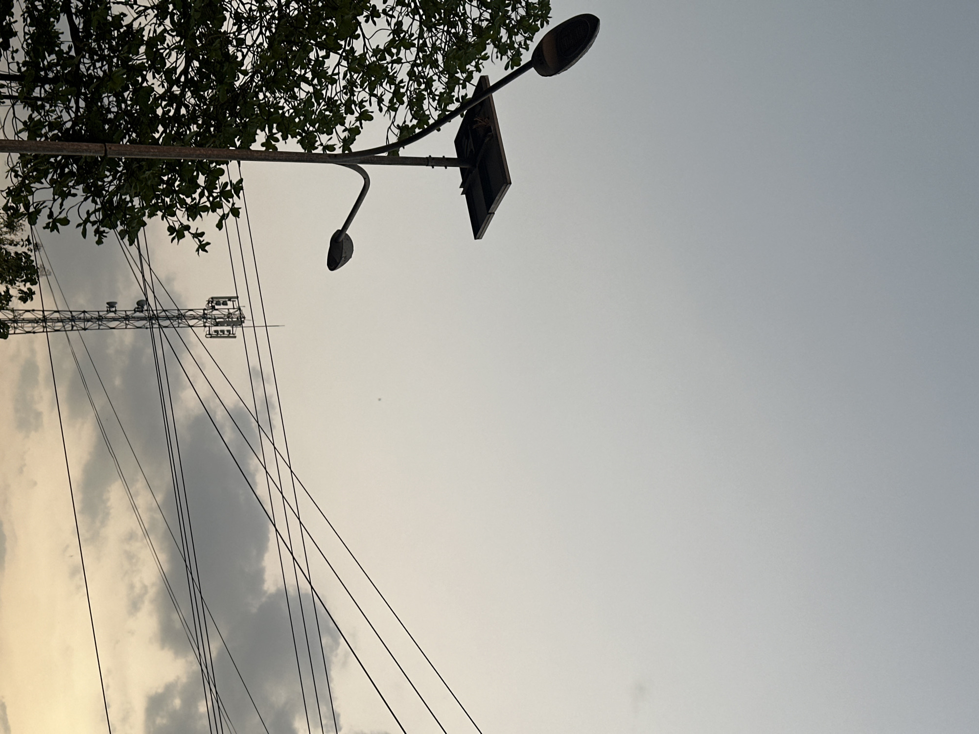 This image depicts a street scene in Bulakrejo, Indonesia, captured from a low angle looking upwards towards the sky. The foreground features a street lamp with a solar panel attached to its pole. The lamp has two lights, one pointing upwards and another angled downwards. To the left of the street lamp, a portion of a leafy green tree is visible. In the mid-ground, a cellular tower stands tall, its lattice structure clearly defined against the sky. Several power lines crisscross the frame, extending from the right side and disappearing towards the left. The sky above is overcast with a mix of light gray clouds and a pale, hazy yellow glow near the horizon, suggesting either sunrise or sunset. The overall atmosphere is muted and somewhat diffused due to the cloud cover. There are no people visible in the image.