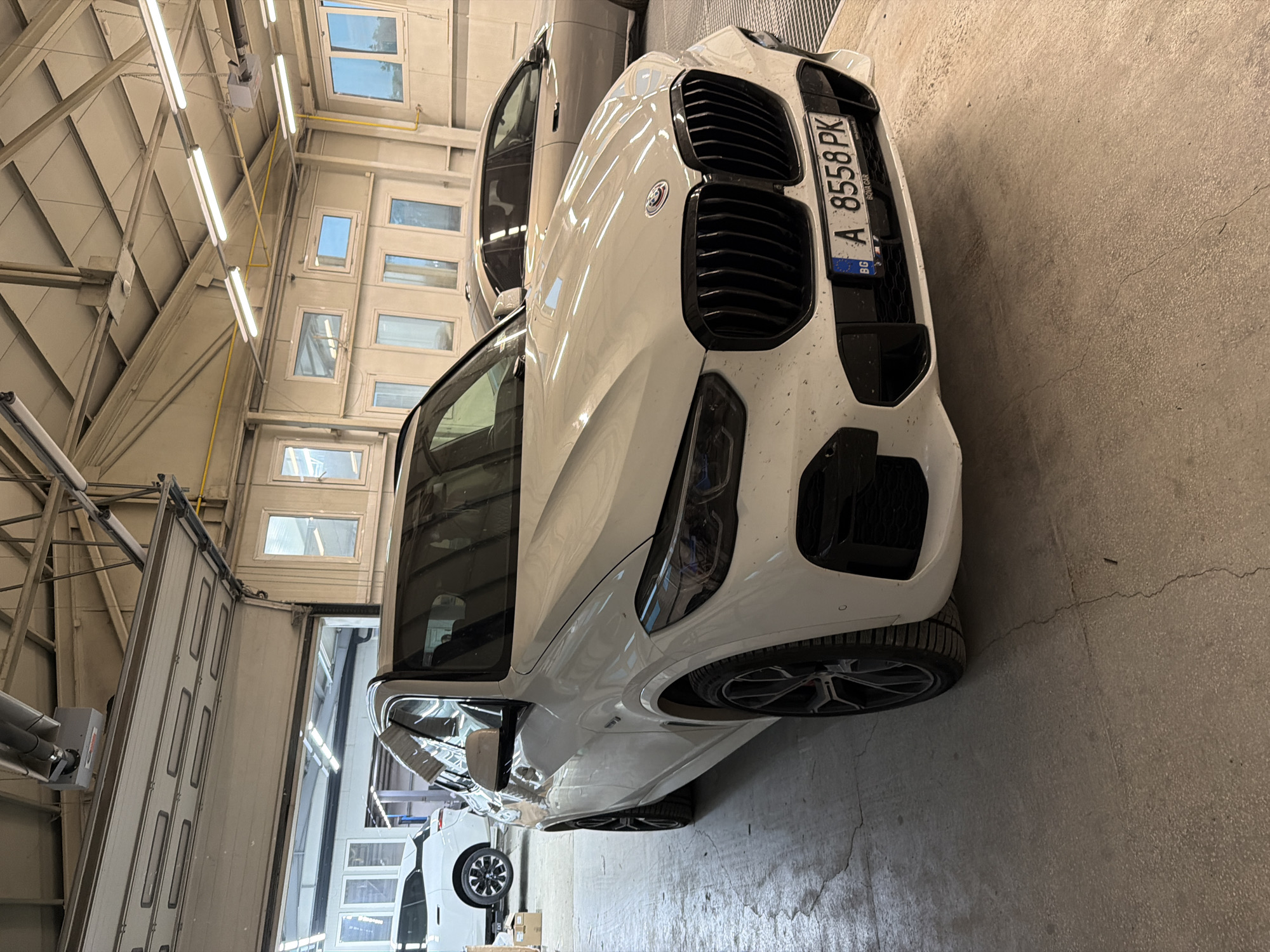A white modern SUV-coupe, likely a BMW X6 or similar model, is parked indoors in what appears to be a well-lit garage or workshop. The car is angled towards the viewer, showcasing its front with large black kidney grilles and an Alpina badge on the grille. A Bulgarian license plate, "A 8558 PK," is prominently displayed on the front, with "BG" indicating Bulgaria and "BOYA CAR" faintly visible below the number. The 'A' prefix confirms the registration location as Burgas, Bulgaria.
The floor is made of light grey concrete, and the background features an industrial-style ceiling with numerous fluorescent tube lights and metal structural beams. Several high-set windows on the wall suggest it is daytime. To the left, a large, white sectional garage door is visible, partially open. Another light-colored car is partially visible behind the primary white vehicle, and a third white vehicle is seen further back in the facility. No people are visible, and no specific activity is taking place; the scene depicts parked cars in an indoor environment.