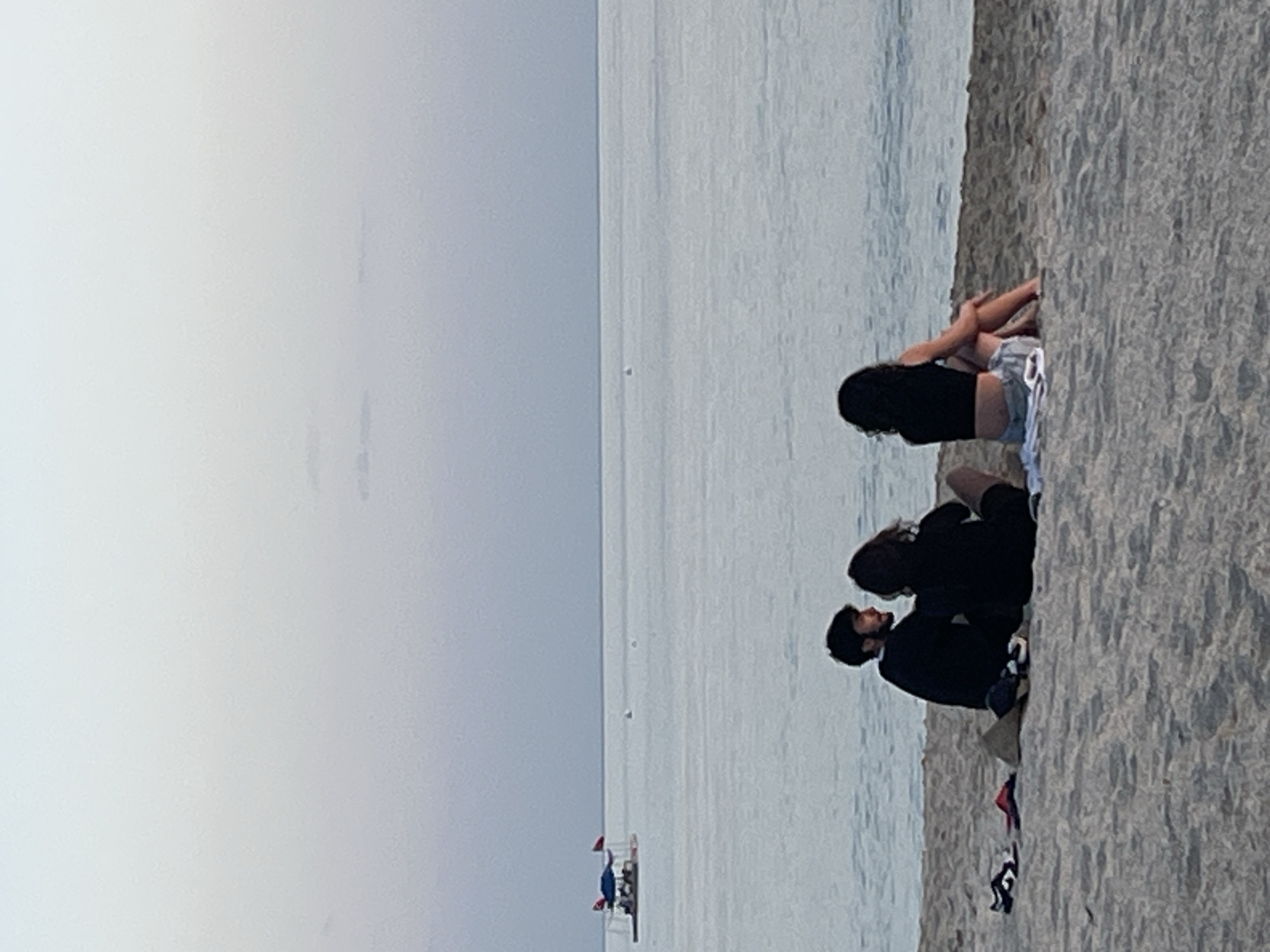 The image depicts a beach scene in Alanya, Turkey. Three individuals are seated on the sand along the shoreline. One person is sitting alone facing the water while the other two are sitting together. The sea is calm, with a partially visible structure that appears to be a boat or raft with red flags in the distance. The sky is overcast.
