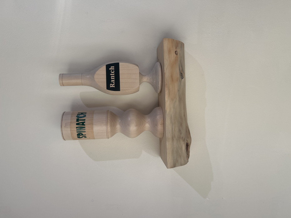 The image displays two decorative, light-colored wooden objects positioned on a rustic wooden shelf, which is mounted on a plain, light-colored wall in Los Angeles, United States of America. The wall exhibits minor texture variations and faint shadows cast by the objects and shelf, indicating a light source from the upper left.
The shelf itself is a single piece of live-edge wood, natural in tone with visible grain and slight imperfections, securely affixed horizontally to the wall.
On the left, a tall, turned wooden object resembles a goblet or vase with a narrow top, a wider cylindrical upper body, an hourglass-shaped stem, and a flat base. The word "SPINATCH" is printed in dark green, capital letters on the cylindrical section of its body.
On the right, a shorter, turned wooden object resembles a stylized bottle with a narrow neck, rounded shoulders, an oval-shaped body, and a flared base. A rectangular black label is affixed to the widest part of its body, bearing the word "Rantch" in white capital letters.
No individuals or active interactions are present in the scene.