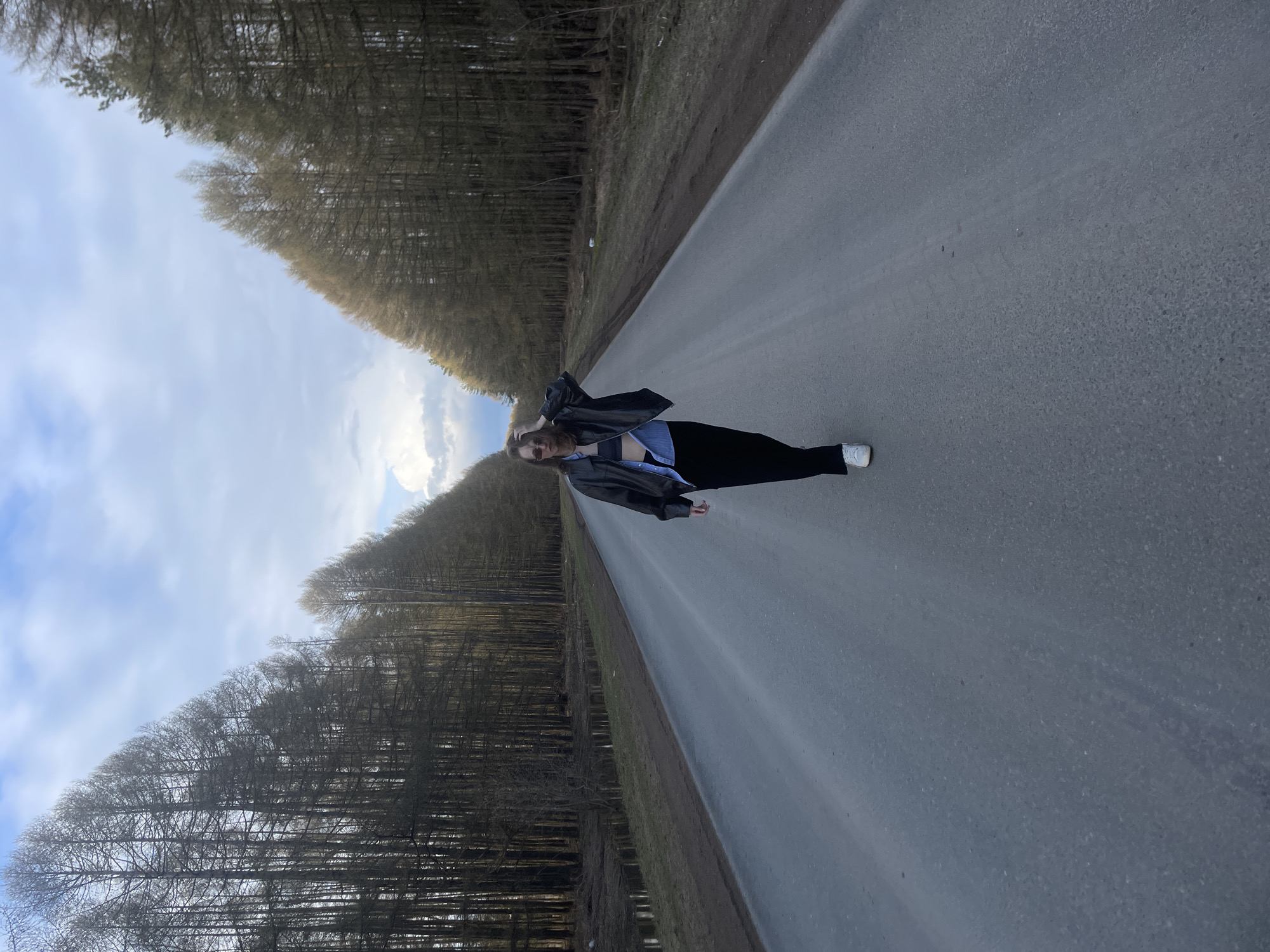 The image displays a full-body shot of a person standing on a paved road, flanked by dense rows of bare trees that recede into the distance. The sky above is partly cloudy with areas of bright blue.
The subject is a young person, likely female, dressed in a black leather jacket over a light blue top, black pants or leggings, and white sneakers. Her long, light-colored hair is visible, and she looks directly at the camera with a neutral expression. One arm is bent with her hand near her head, while the other rests by her side.
The setting is a rural or less-trafficked road cutting through a wooded area. The trees are devoid of leaves, suggesting a late winter or early spring season. The light appears to be diffused, consistent with an overcast day or possibly late afternoon. The scene evokes a sense of solitude, with no other individuals or vehicles present. The asphalt road surface shows minor wear. This natural environment aligns with the location context of Podshivalovo, Russia. No visible text is present in the image.