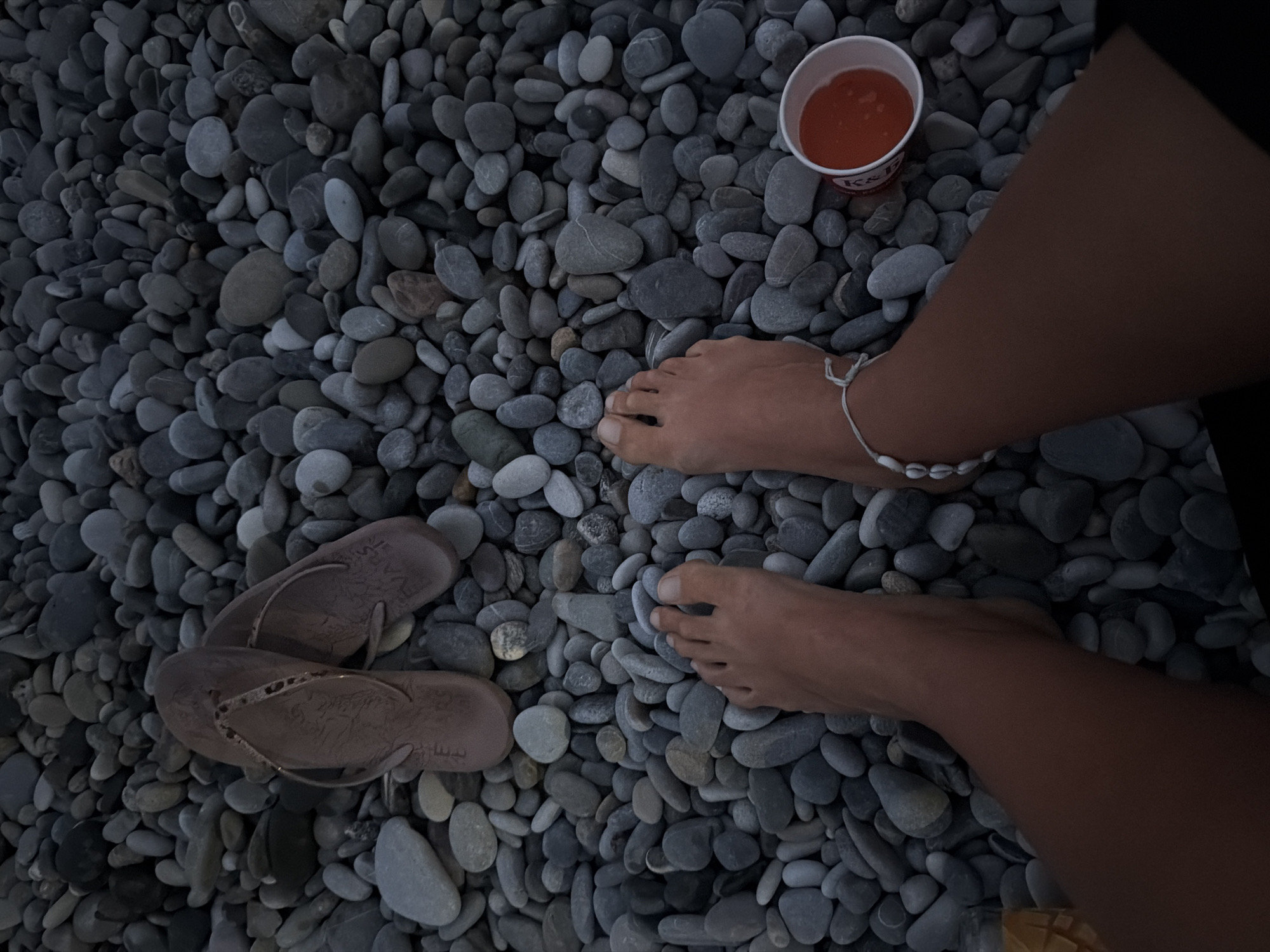 The image depicts a close-up view of a person's bare feet resting on a pebble beach. The pebbles are predominantly gray and smooth, filling the frame. To the left of the feet, a pair of taupe flip-flops with a patterned strap are visible. To the right of the feet, a small white paper cup contains an orange liquid, likely a beverage. The person is wearing a white anklet adorned with small seashells. The lighting suggests it is either early morning or late evening. The scene is consistent with a location on the pebble beaches of Sochi, Russia, which are known for their abundance of smooth, gray stones.