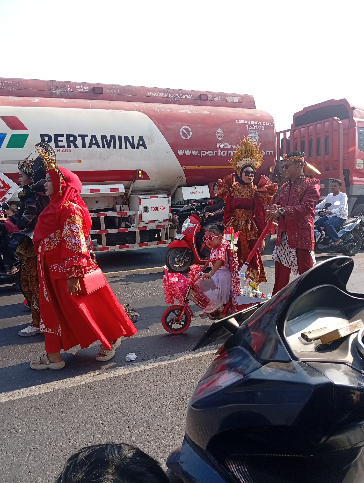 In Sarang, Indonesia, a parade is underway on a sunny day. A large red and white Pertamina fuel tanker truck is parked on the side of the road, serving as a backdrop for the procession. Several individuals are participating in the parade, dressed in vibrant traditional attire. A woman in a striking red hijab and matching dress, adorned with ornate embroidery, is walking prominently in the foreground, carrying a red handbag. Beside her, a man in a matching red outfit with a traditional headdress and sunglasses smiles. A young girl, also in festive attire and wearing pink sunglasses, sits in a pink bicycle decorated with ribbons. Other participants, including a woman in a dark, elaborate costume, are visible further down the road. Motorcycles are present, with one ridden by a person in a white shirt and another partially obscuring the foreground. The road surface is asphalt, marked with white lines, and the environment appears to be an urban or semi-urban setting.