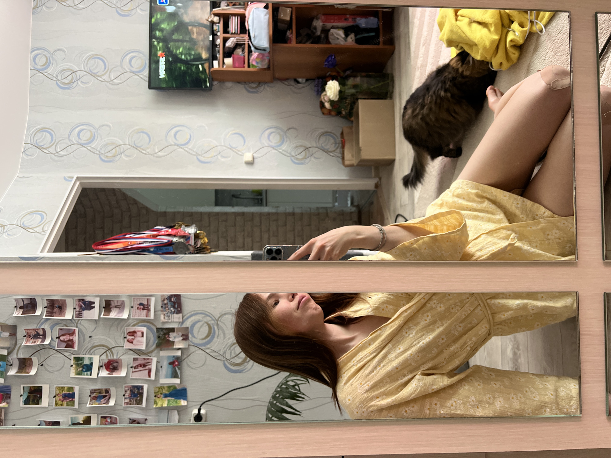 A fragmented view through a set of mirrors captures a scene inside a home, possibly in Chaikovsky, Russia. The reflection shows a woman wearing a yellow floral dress, taking a selfie with a smartphone. In the top portion of the mirror is a dark-furred cat near a yellow garment on the floor. In the background is a television mounted on a wall with patterned wallpaper, a shelf unit, and a floral arrangement. Another mirror shows photos attached to a wall. A racing car toy is above the mirror with the photos.