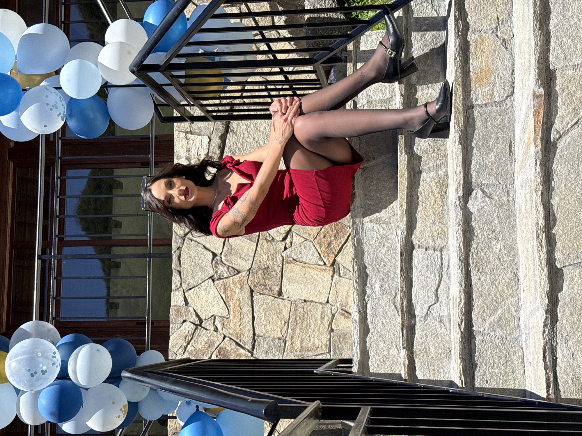 A woman is sitting on an outdoor staircase with a metal railing, amidst decorations of blue and white balloons. The location is Ribaritsa, Bulgaria. The woman is wearing a red dress and black stockings, and black heels. She is facing the camera and has her hands clasped together. The staircase is made of stone, and the background includes a wall with similar stonework and a building with window frames. The balloons are clustered near the staircase and appear to be part of a celebratory event.