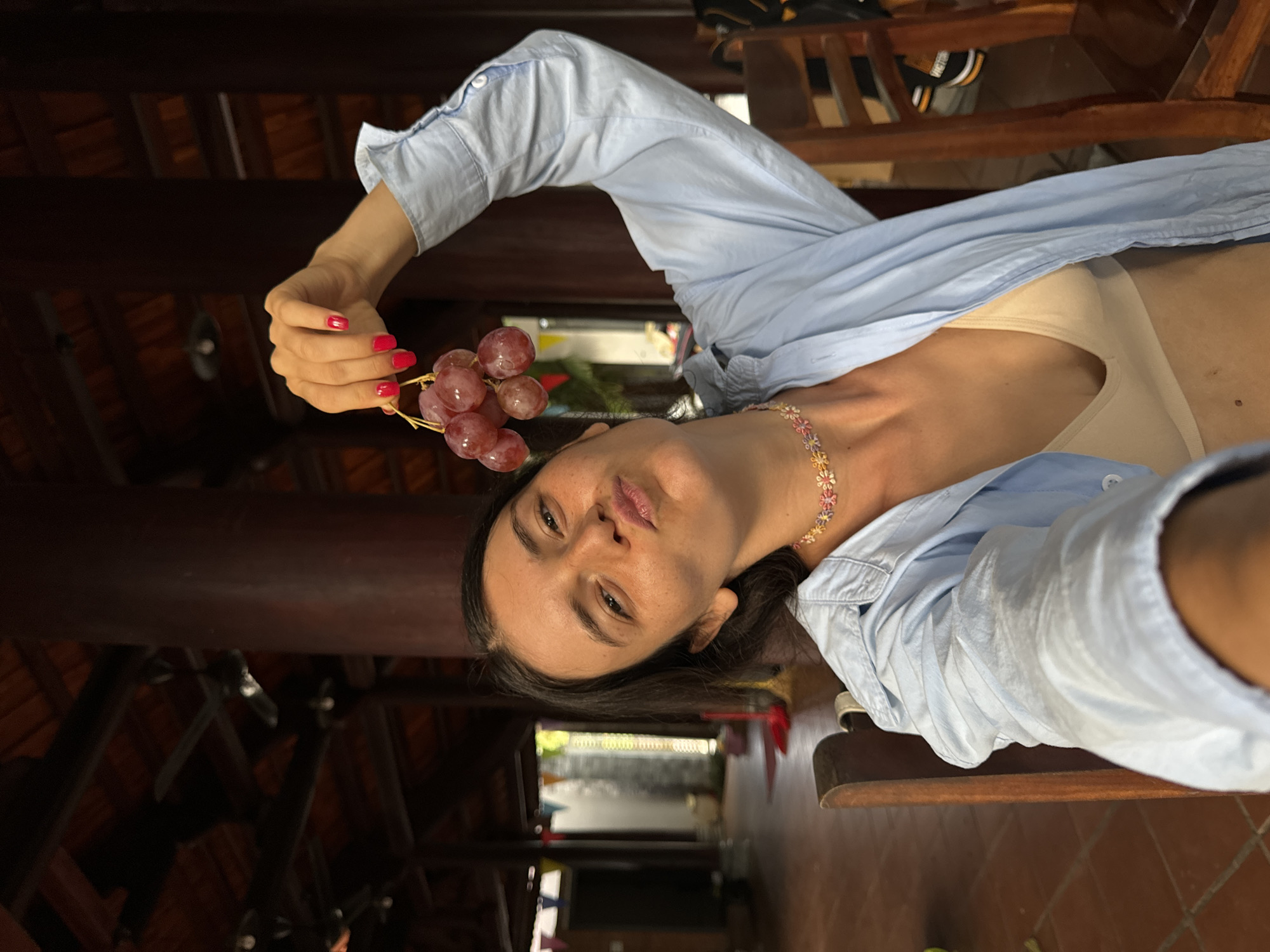 In Mũi Né, Vietnam, a woman with dark hair and light-colored skin is taking a selfie while sitting in a room with a traditional thatched roof and wooden beams. She is wearing a light blue unbuttoned shirt over a tan bralette and a colorful floral necklace. Her right arm is raised, holding a small bunch of red grapes above her head. Her face is turned towards the camera, with her lips pursed in a playful expression and her eyes looking forward. The background shows wooden pillars, ceiling fans, and a glimpse of an outdoor area with colorful flags. The overall scene suggests a casual and relaxed atmosphere.