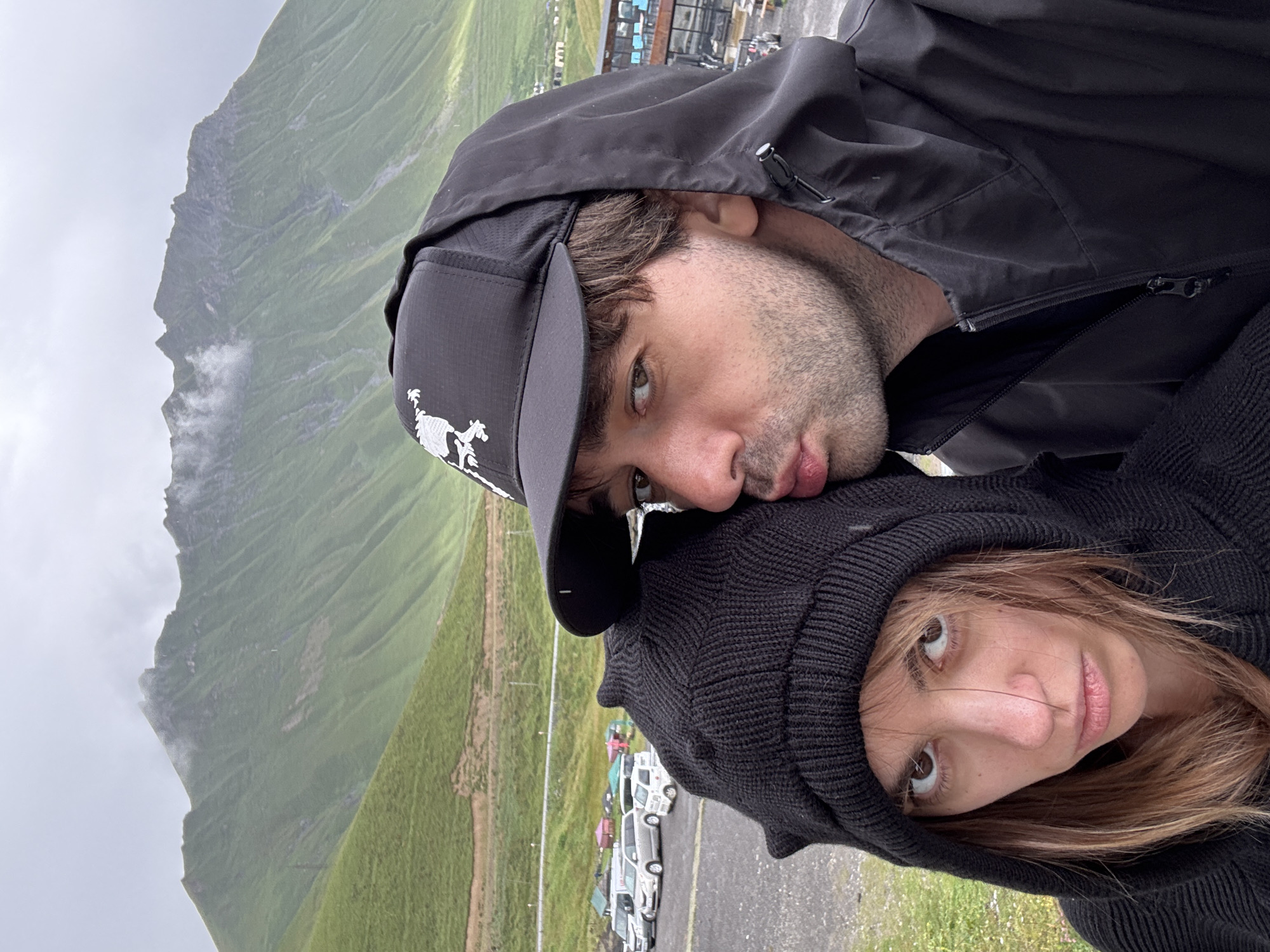 In Gudauri, Georgia, a couple is captured in a close-up selfie against a backdrop of green mountains. The man, wearing a black baseball cap and a black hooded jacket, is puckering his lips towards the woman. The woman, with a black knit beanie and hood, looks at the camera with a displeased expression. In the background, several cars are parked in a lot, and a few colorful tents are visible. The sky is overcast, and the mountain slopes are covered in lush green grass.