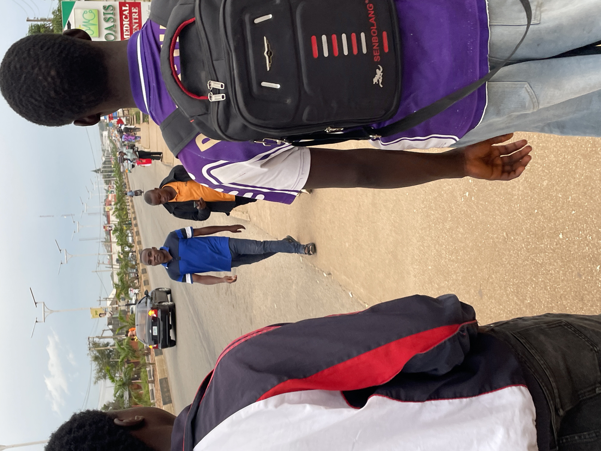 The image captures a street scene in Jos, Nigeria, likely along a busy road that leads into a more commercial or medical area. In the foreground, the backs of two individuals are visible. The person on the left is wearing a white, black, and red jacket over dark jeans. The person on the right is wearing a purple t-shirt with white accents and light blue jeans, carrying a black backpack with the brand name "SENBOLANG" visible.
Further down the road, a man in a blue and black polo shirt and dark jeans walks towards the camera. Behind him, another man in an orange polo shirt and black jacket is walking away from the camera. A black sedan is driving on the road. In the background, there are palm trees, streetlights, buildings, and other pedestrians and vehicles, indicating an urban environment. A prominent sign with "OASIS MEDICAL CENTRE" written on it is visible on the right side of the road. The sky is overcast. The overall scene depicts everyday life and movement in a Nigerian city.