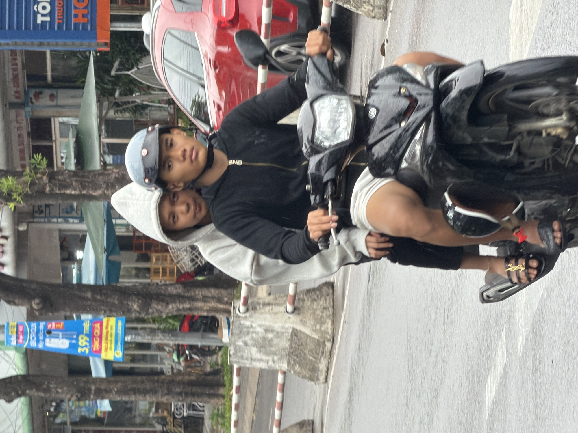 Two young males are riding on a black scooter on a wet street, likely in Nha Trang, Vietnam, as indicated by the signage in Vietnamese. The front rider wears a black zip-up hoodie, white shorts, and a grey helmet. The passenger behind him, wearing a white hooded sweatshirt, appears to be looking towards the camera. A red car is visible in the background. The street is lined with trees and shops, and traffic barriers are present on the side of the road. A blue sign with Vietnamese text and numerals "3.99" is hanging from a pole.