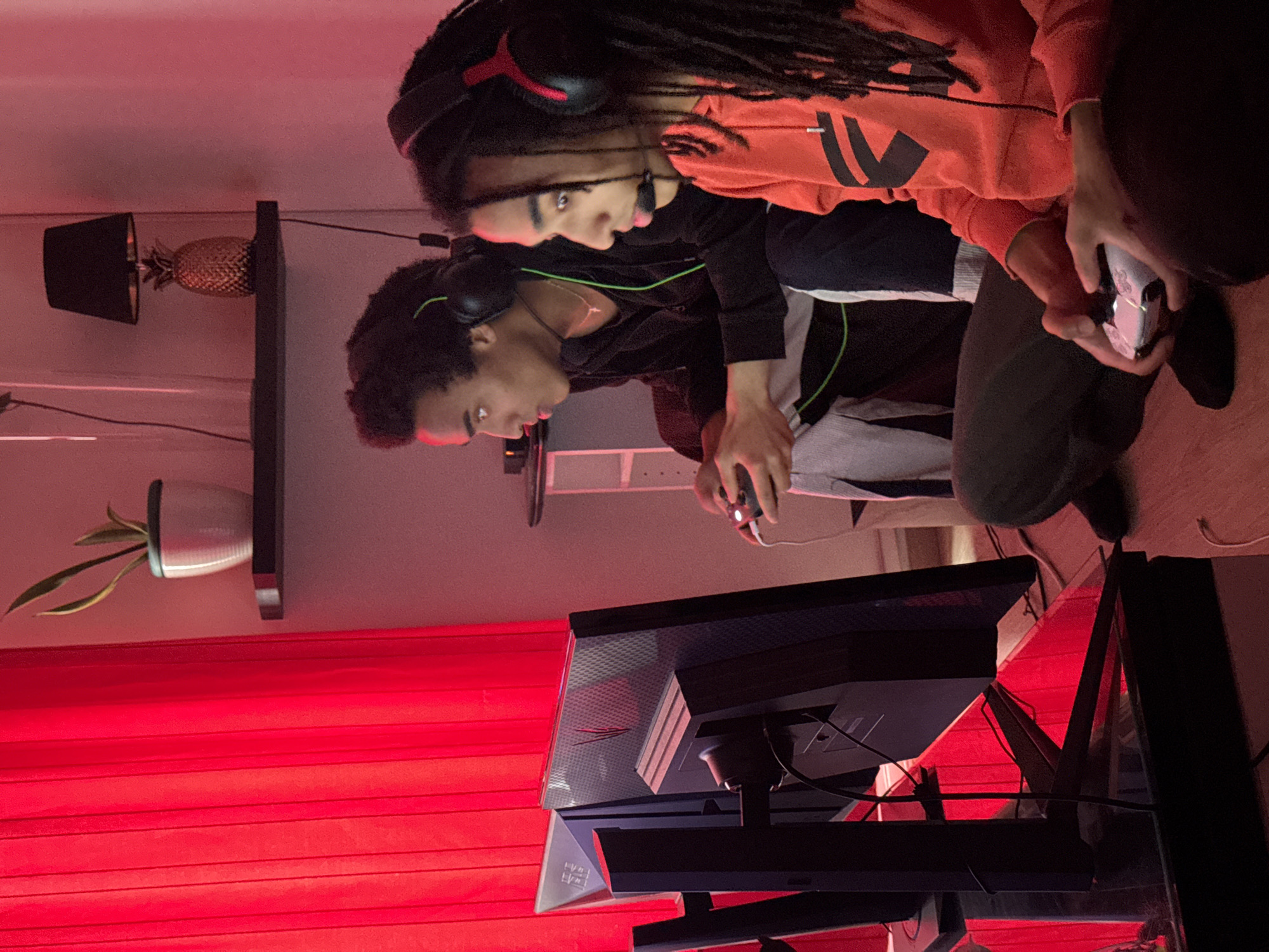 Two young men are playing video games indoors in a room lit with red light. The individual on the left is operating a computer mouse, while the person on the right is holding a gaming console controller. Both individuals are wearing headphones. The setting includes a computer monitor, a shelf with a pineapple-shaped object, and a houseplant. There is no evidence suggesting this image was taken in Paris, France.