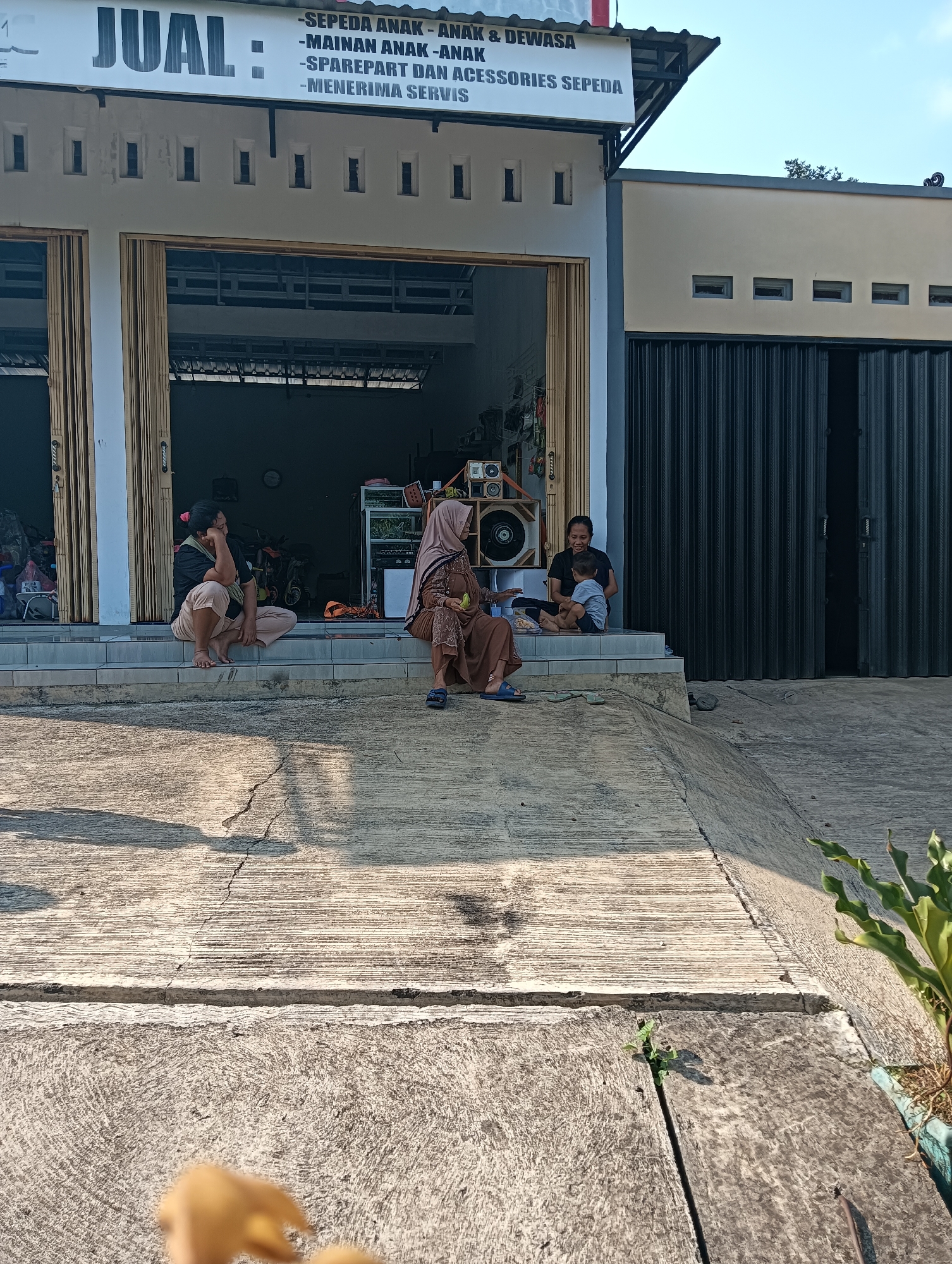 In Cluwak, Indonesia, outside a storefront with a sign that reads "JUAL: -SEPEDA ANAK - ANAK & DEWASA - MAINAN ANAK - ANAK - SPAREPART DAN ACCESSORIES SEPEDA - MENERIMA SERVIS" (which translates to "FOR SALE: -CHILDREN'S & ADULT BIKES - CHILDREN'S TOYS - BIKE SPARE PARTS AND ACCESSORIES - WE ACCEPT SERVICE"), three individuals are seated on the steps in front of the store. To the left, a person in a black t-shirt and light-colored pants sits with their knees bent, resting their chin on their hand. In the center, a woman in a brown hijab and patterned dress sits with her legs crossed, holding a small object in her left hand. To the right, a man in a black t-shirt and dark shorts is seated, holding a young child on his lap who is wearing a light blue shirt and dark shorts. The storefront has a closed metal gate on the right and open sliding doors on the left, revealing items stored inside, including bicycles. The ground in front of the steps is concrete, with some cracks visible. A potted plant is visible in the lower right corner of the frame. The scene appears to be outdoors in daylight, with shadows cast on the concrete.