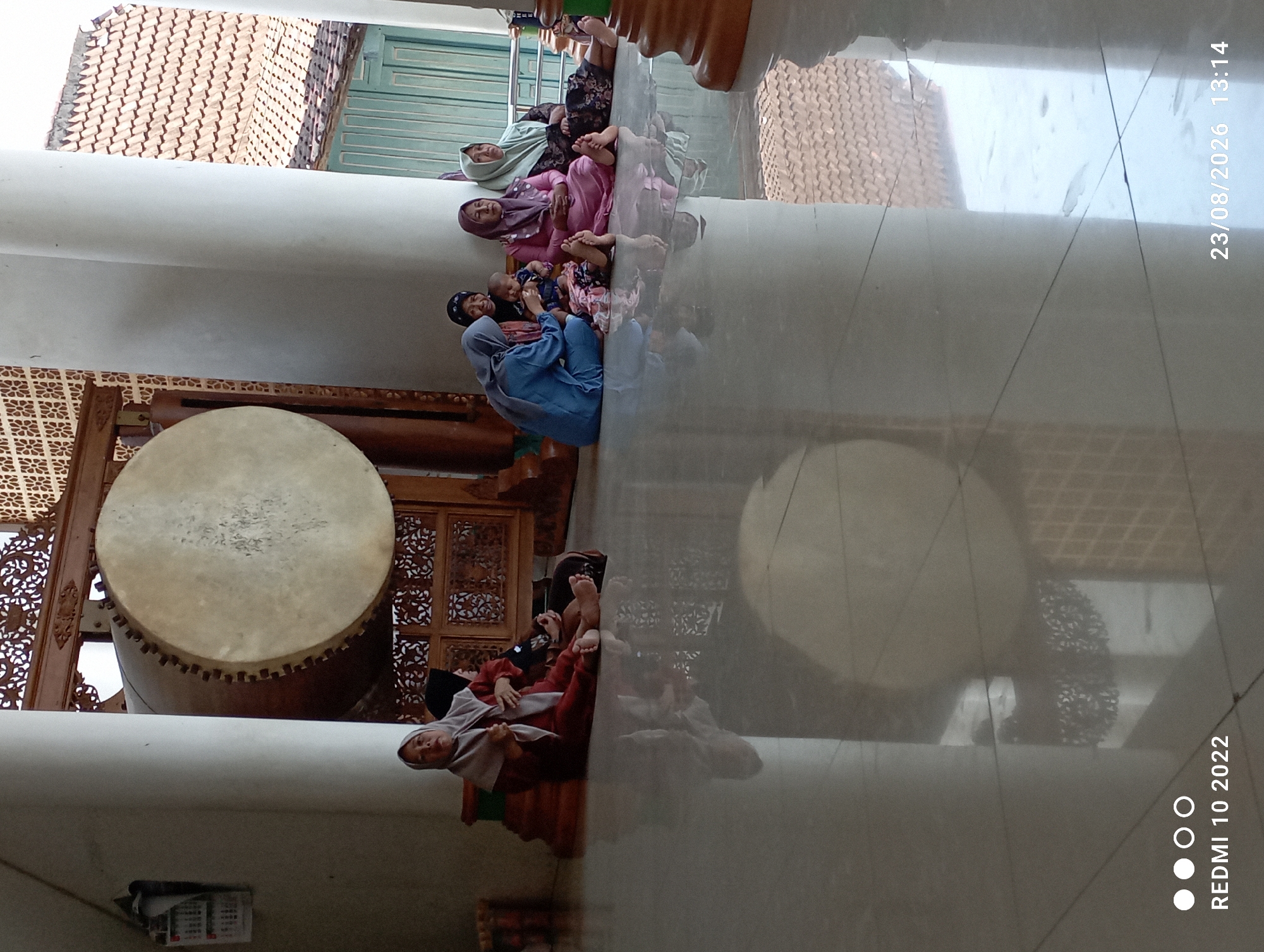 A group of five Muslim women, some with headscarves, sit on the floor of a building with a large drum in the background. The woman on the far left wears a maroon headscarf and dress. Next to her, another woman in a dark headscarf sits with her legs crossed. The third woman from the left is dressed in blue and is holding a baby. The fourth woman wears a purple headscarf and a pink dress, and the woman on the far right has a green headscarf and a patterned dress. The floor is polished and reflects the scene. The building appears to be a place of worship or cultural significance, possibly a mosque or a traditional Indonesian building, given the presence of the large drum and the architectural style. The overall setting suggests a gathering or a moment of rest. The date stamp "23/08/2026 13:14" and "REDMI 10 2022" are overlaid on the image. The location is specified as Dukuhseti, Indonesia.
