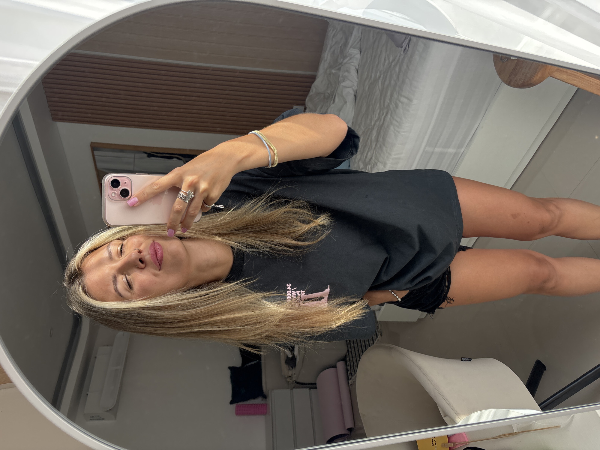 A young woman with blonde hair, wearing a black t-shirt and black shorts, takes a selfie in a mirror. She is wearing rings and bracelets on her hands. The room appears to be a bedroom with a bed, a dresser, and a chair visible in the background. The woman's eyes are closed, and she has a slight smile on her face. The lighting suggests it is daytime.
