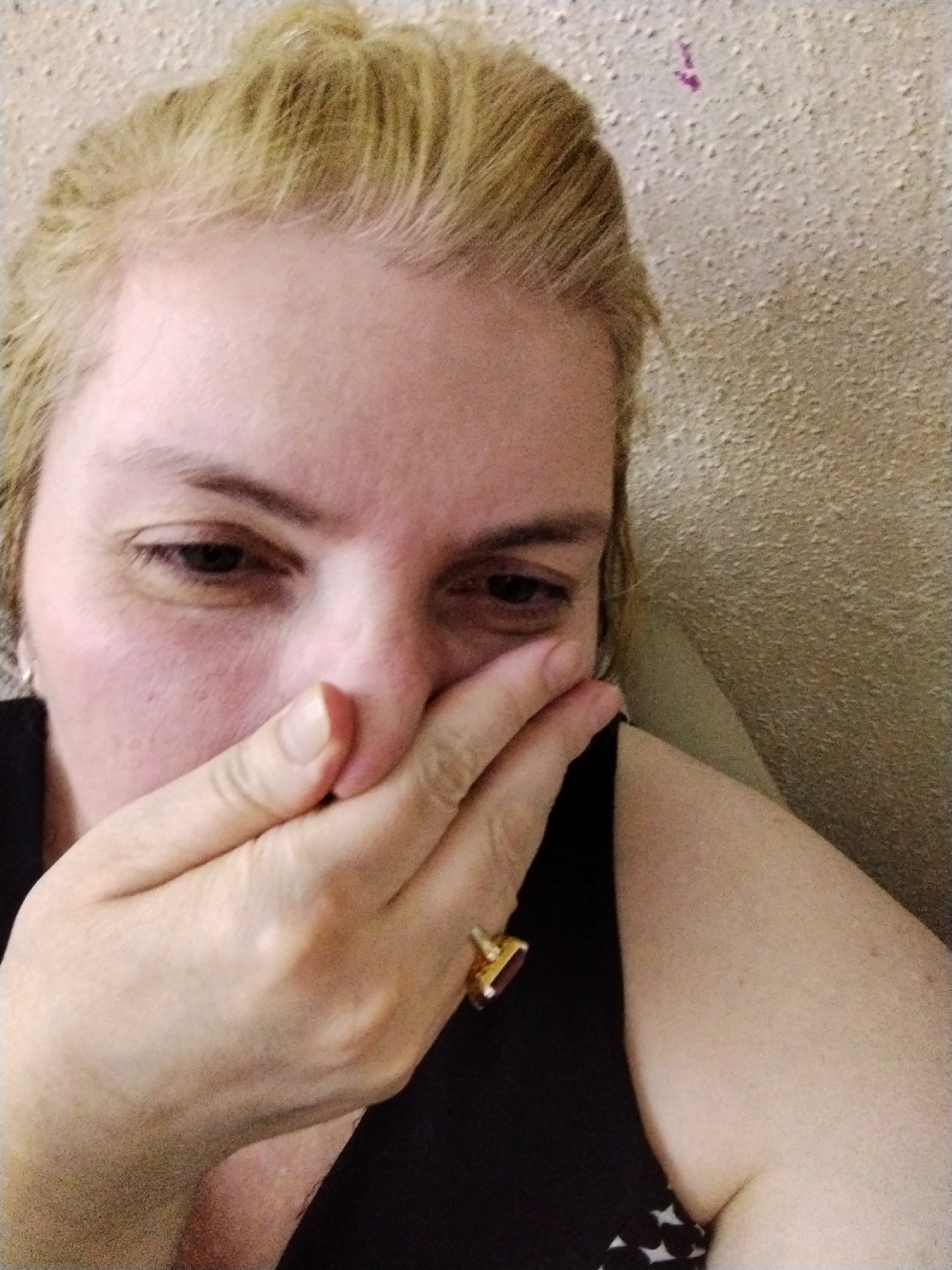 A woman with blond hair is covering her mouth with her right hand. She is wearing a black sleeveless top and a ring with a rectangular stone. The background is a textured beige wall. The location and the city of Al Mas'oudi, United Arab Emirates are not discernible from the image.