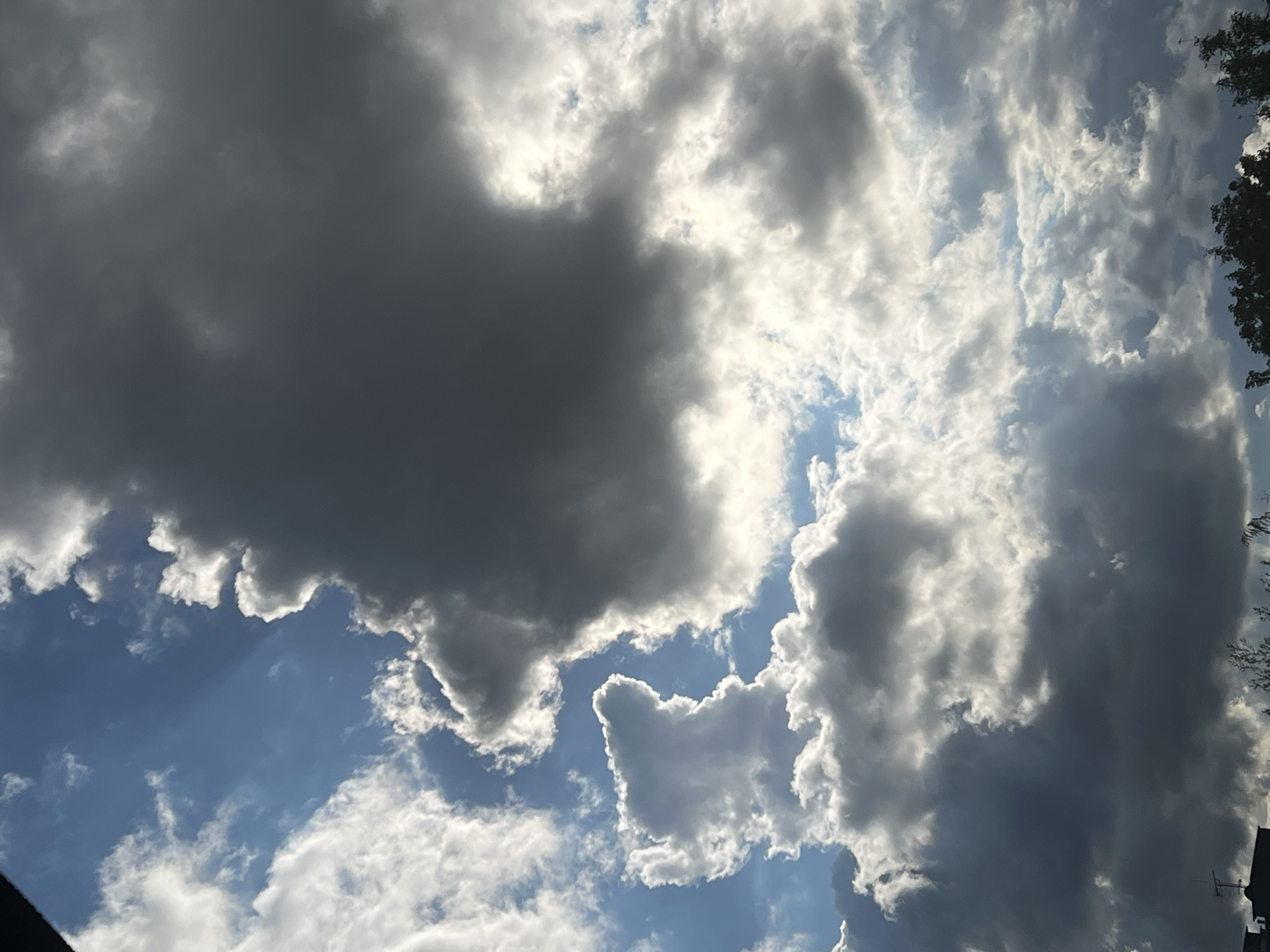 The image depicts a dramatic sky with a mix of dark, imposing storm clouds and bright, luminous cumulus clouds, backlit by the sun. The sky is a vibrant blue in some areas, with sunbeams piercing through the denser cloud formations. At the very bottom of the image, the silhouette of a building with an antenna and the tops of trees are visible, suggesting an urban or suburban setting. The play of light and shadow across the clouds creates a sense of depth and atmospheric intensity. The scene is consistent with a weather event occurring in Warsaw, Poland.