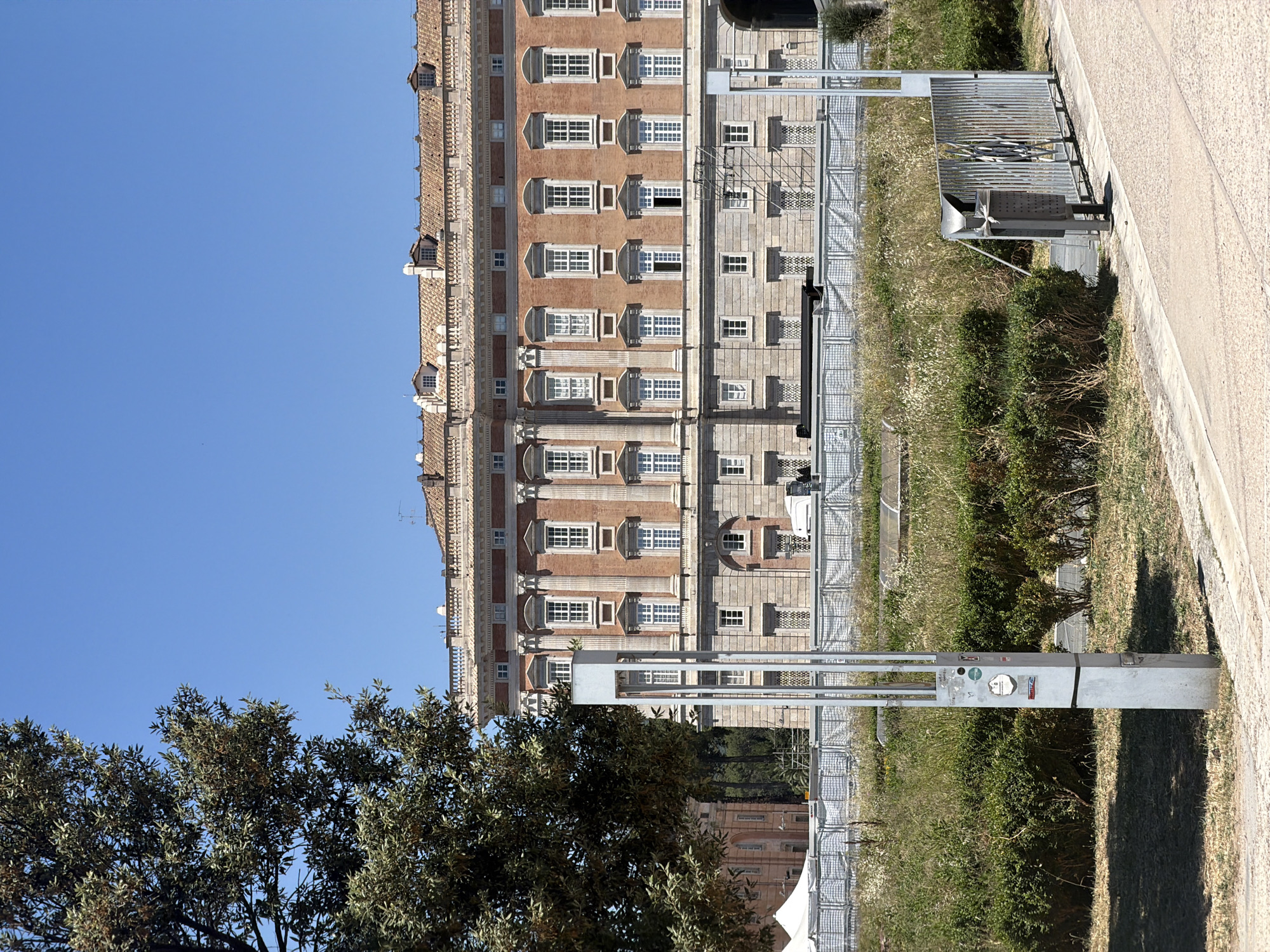 The image shows a side view of the Royal Palace of Caserta, Italy. The facade of the palace is visible on the left, with multiple rows of windows and a light-colored exterior. To the right of the palace is a fenced area with a small structure and vegetation. In the foreground, there's a tree on the left and a paved area on the right. The sky is clear blue.