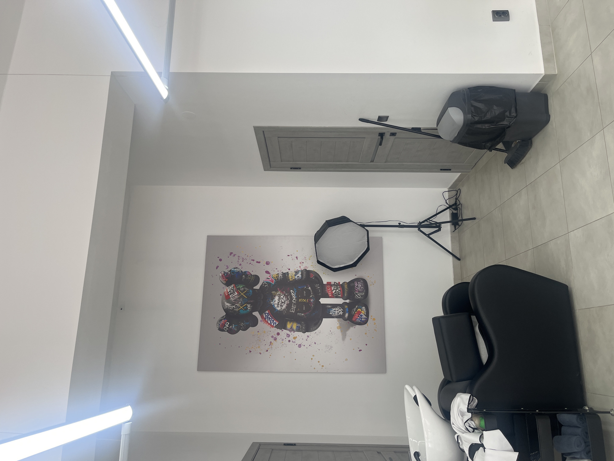 The image depicts an indoor scene, likely a studio or salon, with a contemporary aesthetic. A large, colorful canvas art piece featuring a stylized character is prominently displayed on a white wall. To the left of the artwork, a professional photography light on a tripod is set up. In the foreground, a black salon chair with a headrest is positioned next to a white wash basin, suggesting a hair salon or barber shop. A rolling cart with towels and other salon supplies is visible next to the chair. To the right of the artwork, a closed gray door with horizontal paneling stands against the wall. Next to the door, a black trash bin with a gray lid is placed on the floor, accompanied by a broom. The room is illuminated by long, linear fluorescent lights mounted on the ceiling. The floor is tiled with large, light gray square tiles. The walls are painted a neutral white. There are no people visible in the image. The scene does not contain any specific geographical markers that would allow for identification of the city of Uzhhorod, Ukraine.