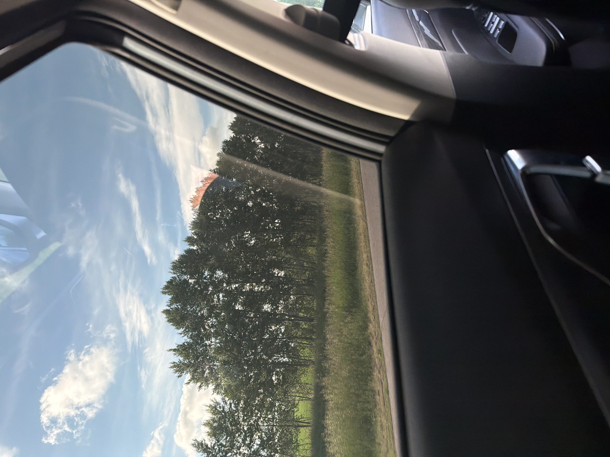This image captures a view from inside a vehicle, looking out through its side window onto a bright, sunny landscape. The scene is dominated by a clear blue sky adorned with scattered white clouds, ranging from wispy cirrus to more defined cumulus formations. Below the sky, a dense line of tall, leafy green trees extends across the mid-ground, indicating a wooded area or forest edge. A lush green field or grassy verge stretches from the base of the trees, meeting what appears to be a narrow, dark road or pathway along the bottom edge of the exterior view.

The dark frame of the car window and parts of the vehicle's interior, including a door panel and hints of other components like a door handle or dashboard details, frame the outdoor scene. Reflections of the sky and clouds are visible on the window glass, subtly overlaying the distant landscape. The time of day is clearly daytime, characterized by ample sunlight, which creates bright highlights and some contrast in the foliage.

No people are visible in the image, and there is no discernible text. The overall impression is that of a peaceful drive through a rural or natural environment, likely in the area of Akkuzovo, Russia, given the provided context. The weather is fair, with bright conditions and a blue sky.