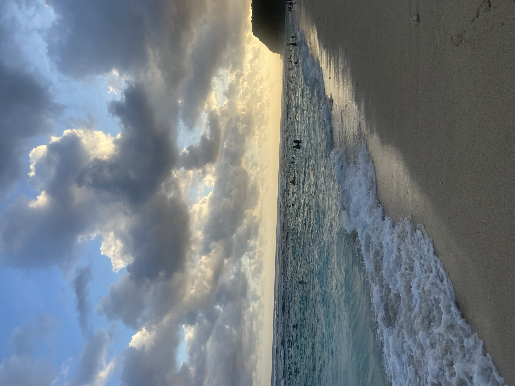 The image shows a beach scene in Kuta Selatan, Indonesia. The beach is sandy and there are waves breaking on the shore. Several people are swimming in the ocean. The sky is cloudy with a patch of sunlight shining through. The sun is positioned on the horizon line, creating a glow in the sky. There is a mountain or cliff in the background.