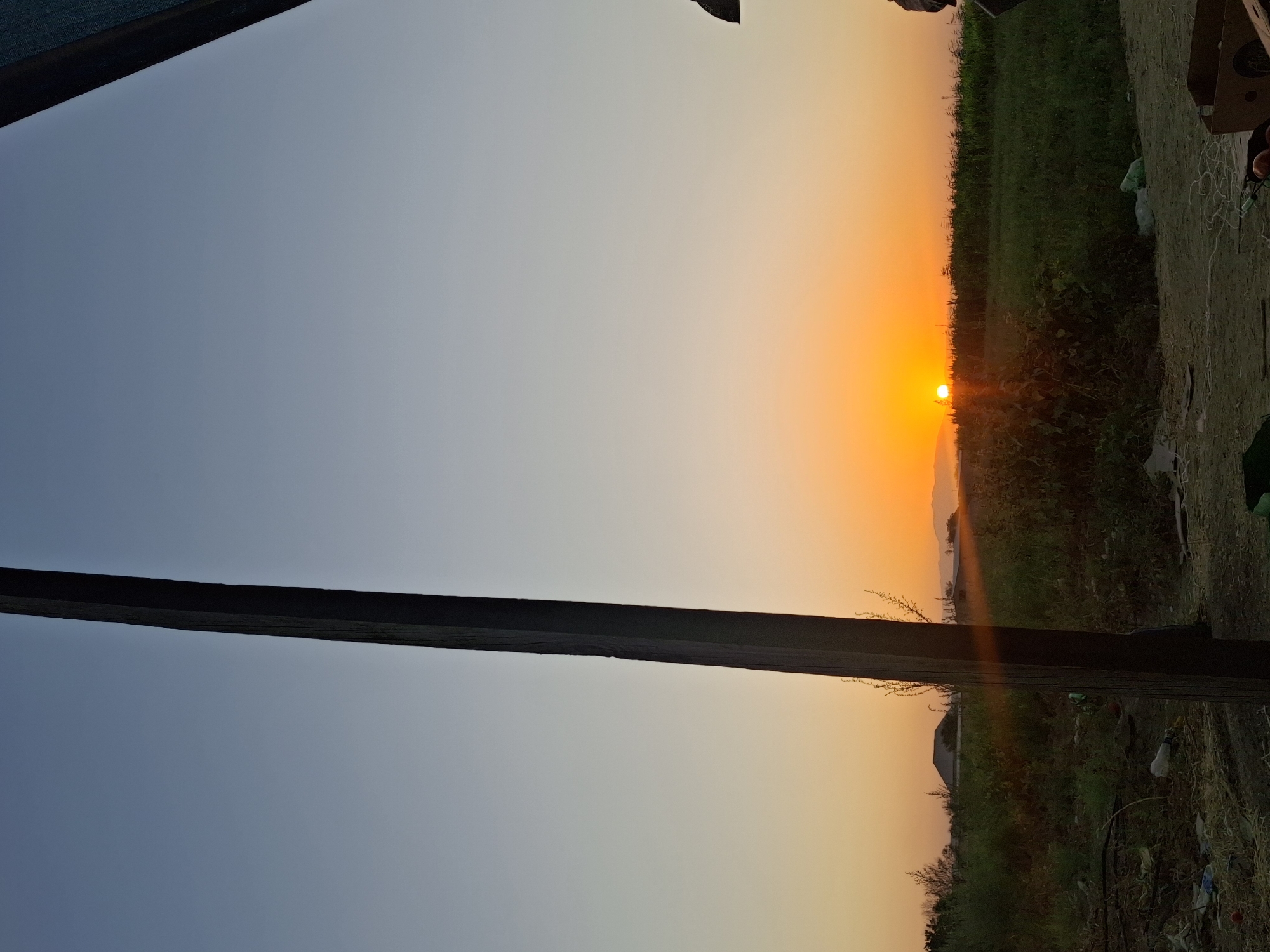 The image captures a sunset over a rural area, likely in or near Utmayli, Uzbekistan. The sky transitions from a pale blue at the top to a vibrant orange glow near the horizon where the sun is setting. A dark, vertical wooden post is prominent in the foreground, dividing the frame. To the left of the post, sparse vegetation and some scattered debris, including plastic bags and wrappers, are visible on the ground. Beyond this, a field of green grass and plants stretches towards the horizon, with a building or structure visible in the distance. The right side of the image shows a dark, indistinct object, possibly part of a structure or canopy. The overall impression is a natural landscape viewed from a somewhat unkempt or undeveloped location.