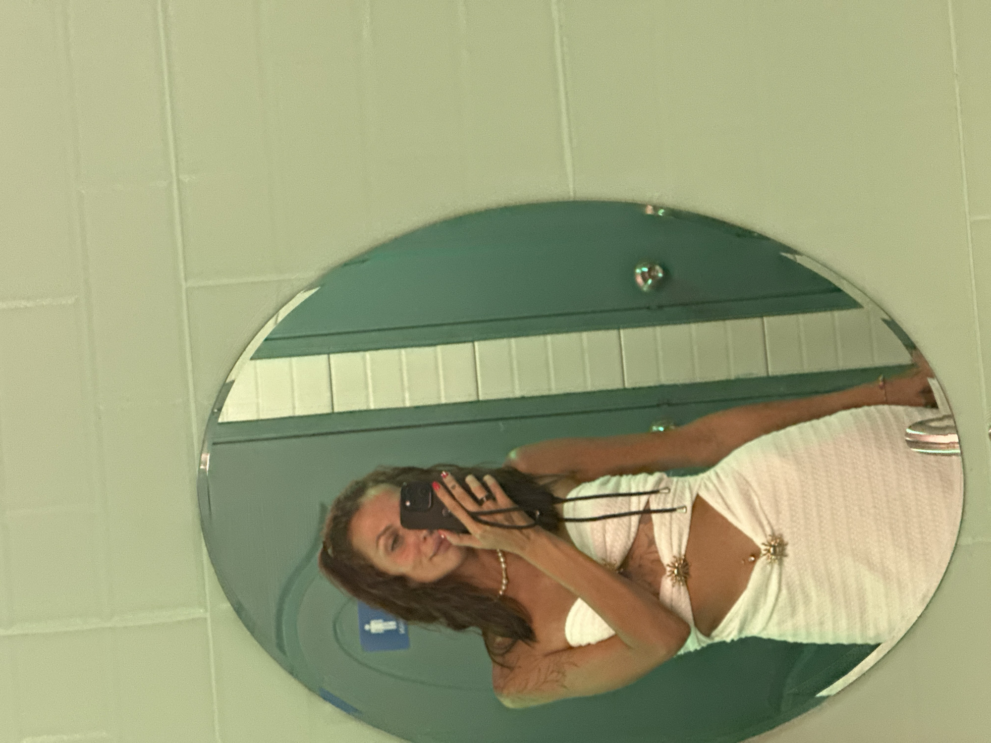 The media file shows a woman taking a selfie in a bathroom mirror. The woman has long, brown hair and is wearing a white, strapless dress with cutouts on the torso, adorned with star-shaped embellishments. She is also wearing a pearl necklace and has red nail polish. She is holding a black smartphone to take the picture, and a tattoo is visible on her arm. The bathroom has pale green tiled walls and a large, oval mirror. Reflections in the mirror show more of the bathroom, including white tiled walls and a male restroom sign. The lighting suggests it is daytime. The overall mood appears casual and confident.