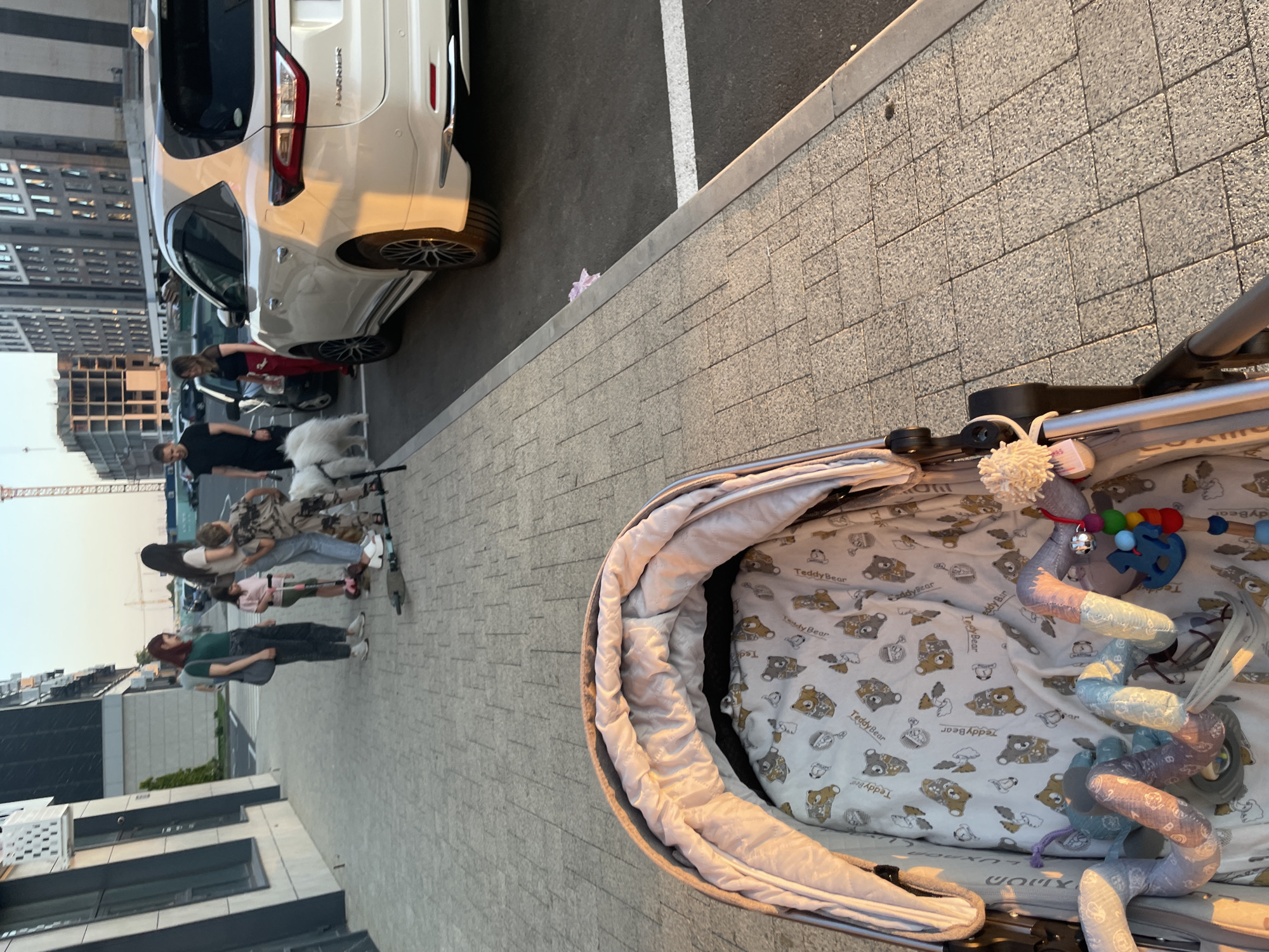 The image depicts a sidewalk scene in Stavropol, Russia, likely near a residential or commercial complex given the modern architecture of the buildings in the background. In the foreground, a baby stroller is positioned, with its interior visible, displaying a teddy bear-themed lining and attached toys.
On the sidewalk further back, several individuals are present. A woman in a green shirt and dark pants stands with a gray tote bag. Next to her, a young girl in a pink shirt and shorts is holding a scooter. Behind them, another woman in a light-colored t-shirt and jeans is observing. A child in a camouflage outfit is standing next to a fluffy white dog, possibly a Samoyed, which appears to be a pet. Two men are also visible in the background, one in a black t-shirt and the other partially obscured.
Adjacent to the sidewalk is a parking area where a white SUV is parked. A pink plastic bag is visible on the asphalt near the curb. The setting appears to be during daylight, with the sun casting a warm glow on the scene, suggesting it might be late afternoon or early evening. The ground is paved with light-colored interlocking bricks. The overall impression is a casual, outdoor moment with a family and their pets.