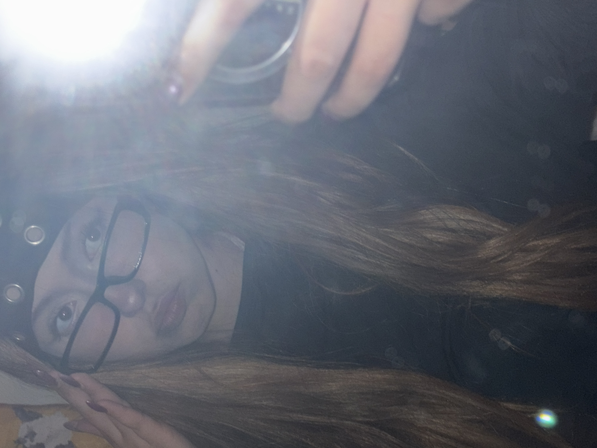 A young person, wearing glasses and a hat with metal grommets, is lying down and taking a selfie with a mobile phone. The flash is on, causing a bright glare at the top of the frame. The background is dark and blurry, but there appear to be some light-colored objects or patterns visible. The person has long brown hair and dark fingernails. There is no evidence to suggest the image was taken in Milan, Italy.