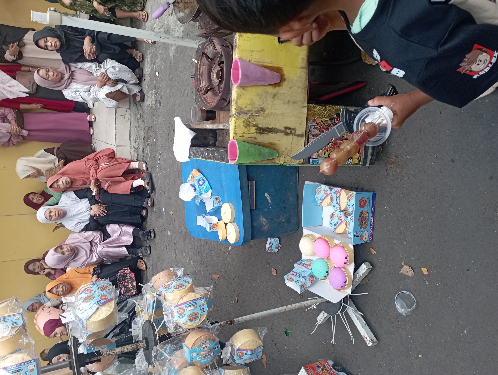 This is a street scene in Tasikmalaya, Indonesia. Several women are sitting and standing near a yellow building. In the foreground, a young boy is holding a skewer of candied fruits and wearing a black t-shirt with "Roblox" logos. A food cart with a stove and colorful cones is to the right of the boy. Various toys, possibly steam buns, are displayed in boxes and bags on a stand. The ground is paved with asphalt, and there are scattered leaves. The overall impression is a bustling outdoor market or street food vending area.