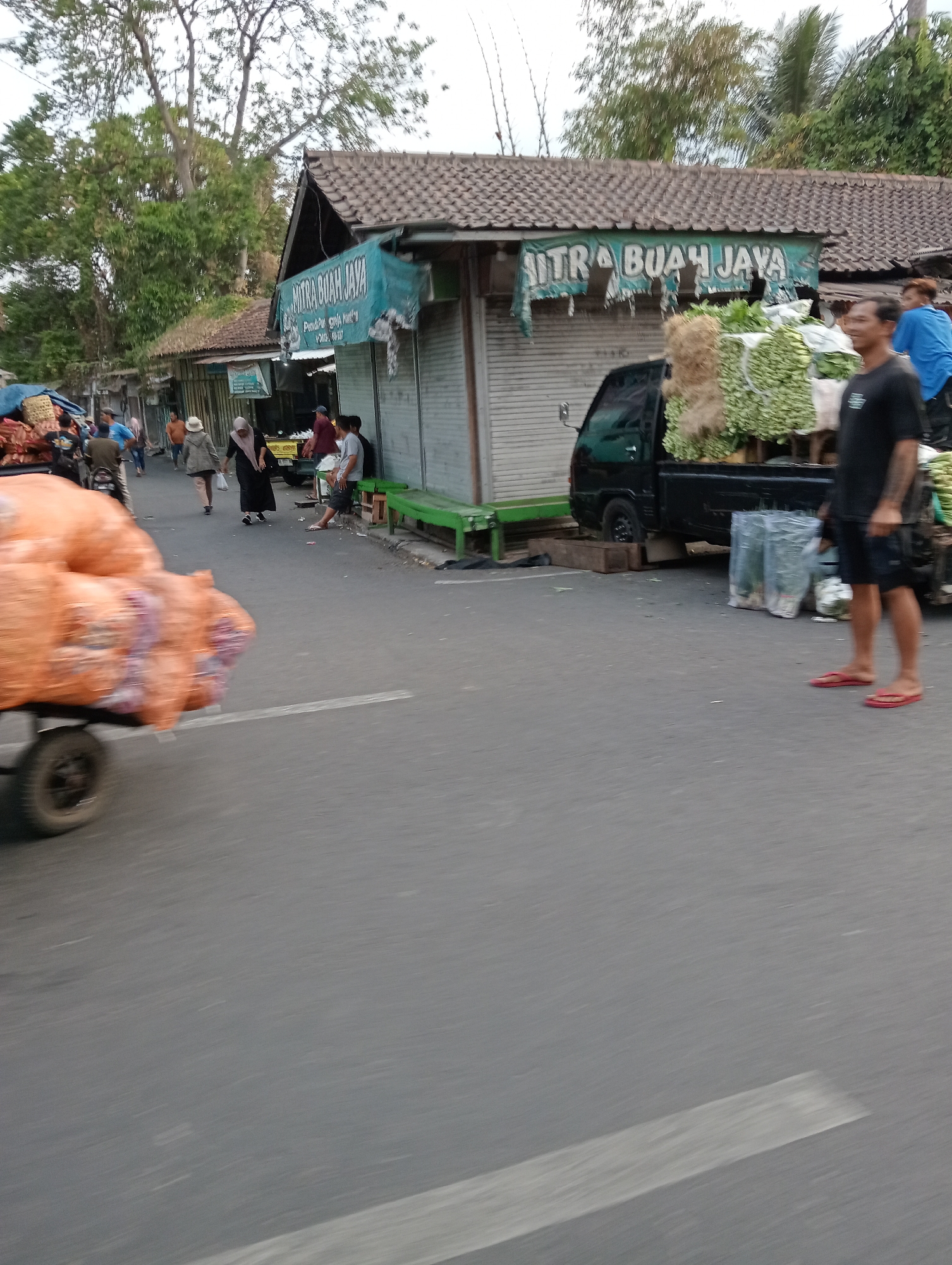 The image depicts a street scene in Muntilan, Indonesia, likely a market area. In the foreground, a handcart laden with large, orange, burlap sacks is being pulled, its movement blurred by motion. To the right, a black pickup truck is parked, carrying a substantial amount of fresh produce, including bunches of leafy greens and what appear to be coconuts or similar fibrous items. A man in a black t-shirt and shorts is standing next to the truck, looking towards the camera, while another man in a blue t-shirt is partially visible behind him. Several plastic bags containing plants or produce are also on the ground near the truck.
Further down the street, several market stalls are visible. One stall has a faded blue banner with "MITRA BUAH JAYA" written on it. People are walking along the street, some appearing to be shoppers or vendors. Among them, a woman in a black hijab and dress walks away from the camera, accompanied by another person. A man in a brown shirt and a woman in a white hat and coat are also visible walking towards the stalls. The stalls themselves have corrugated metal roll-up doors, typical of market shops. Trees line the background, indicating an outdoor setting, and the overall atmosphere suggests a bustling day at a local market. The road surface is asphalt, with a white line painted on it.