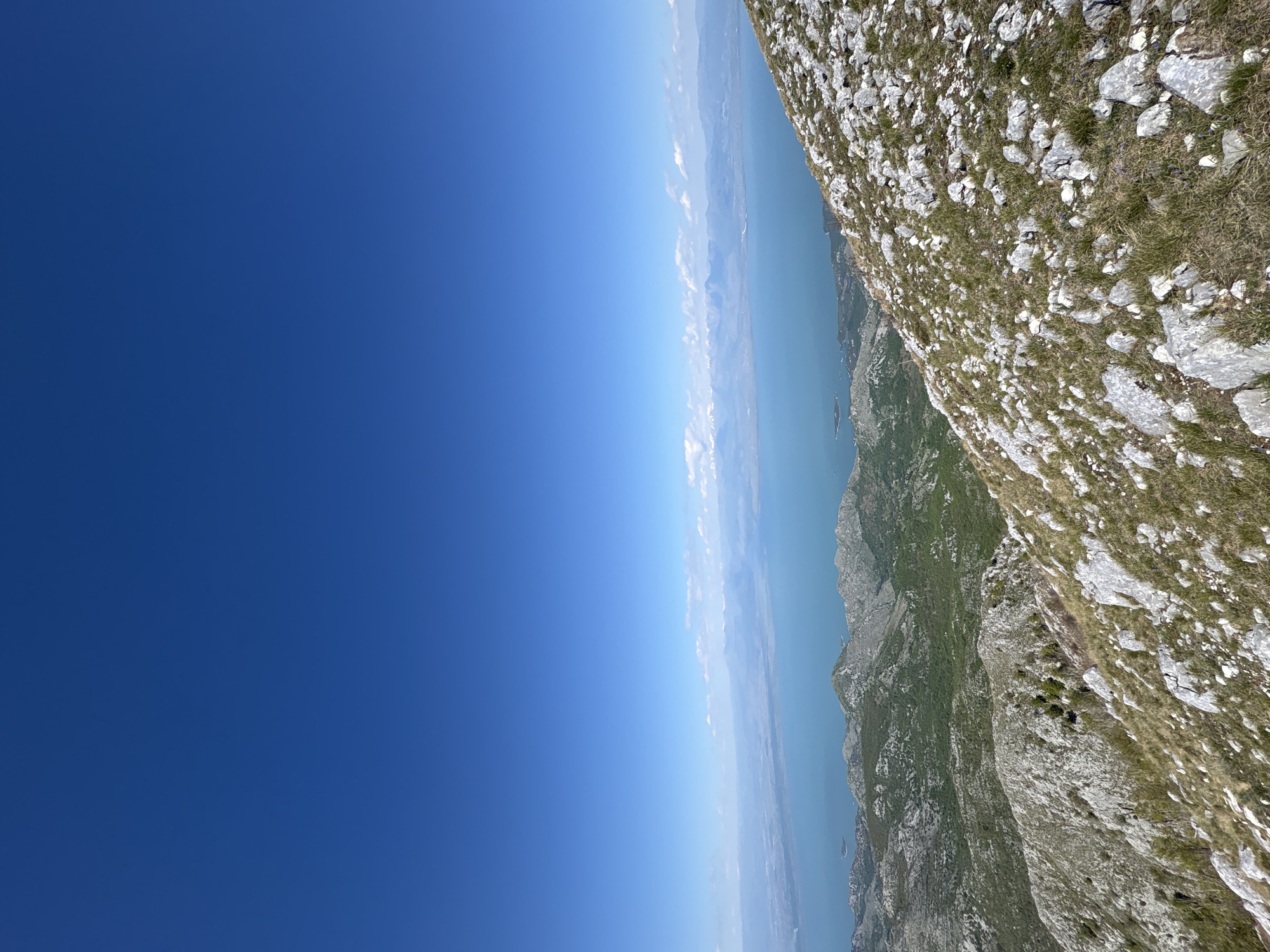 The image shows a mountain landscape in Mali Mikulići, Montenegro, with a view towards the sea. The sky is clear and blue. A rocky mountainside occupies the right side of the frame, with sparse vegetation. In the distance, there is a body of water reflecting the sky, with land faintly visible on the opposite shore.