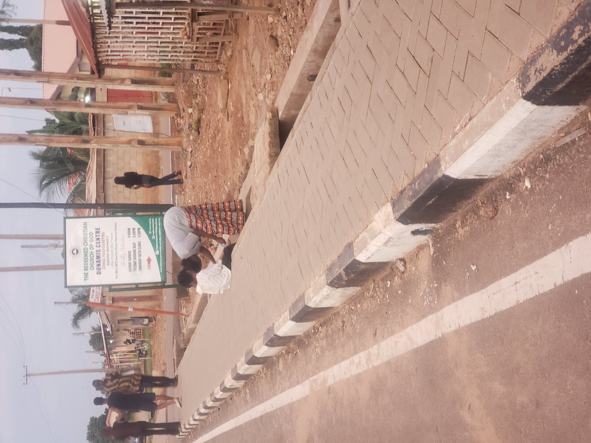 The image captures an outdoor scene on an overcast day, likely in Narayi, Nigeria. In the foreground, a paved walkway runs alongside a road with a white line marking its edge. The walkway has a distinct black and white curb. On the walkway, two individuals are visible. A child in a white patterned shirt sits on the ground, while a person wearing a light-colored top and a patterned skirt is bending down, possibly interacting with the child or the ground.
Further down the walkway, a large sign displays text, including "THE REDEEMED CHRISTIAN CHURCH OF GOD DUNAMIS CENTRE" and information about services and locations in Kaduna Province. Behind the sign, a few people are walking away from the camera, including one person in a black t-shirt and dark shorts, and another in a patterned shirt and dark pants. To the right of the sign, a woman in a black top and dark pants walks away from the camera, and beyond her, there are buildings with corrugated iron roofs and concrete block walls. A small stall with wooden structures and some produce is also visible. The environment appears somewhat dry and dusty, with scattered debris on the ground. Power poles and electrical wires are present in the background, typical of an urban setting.