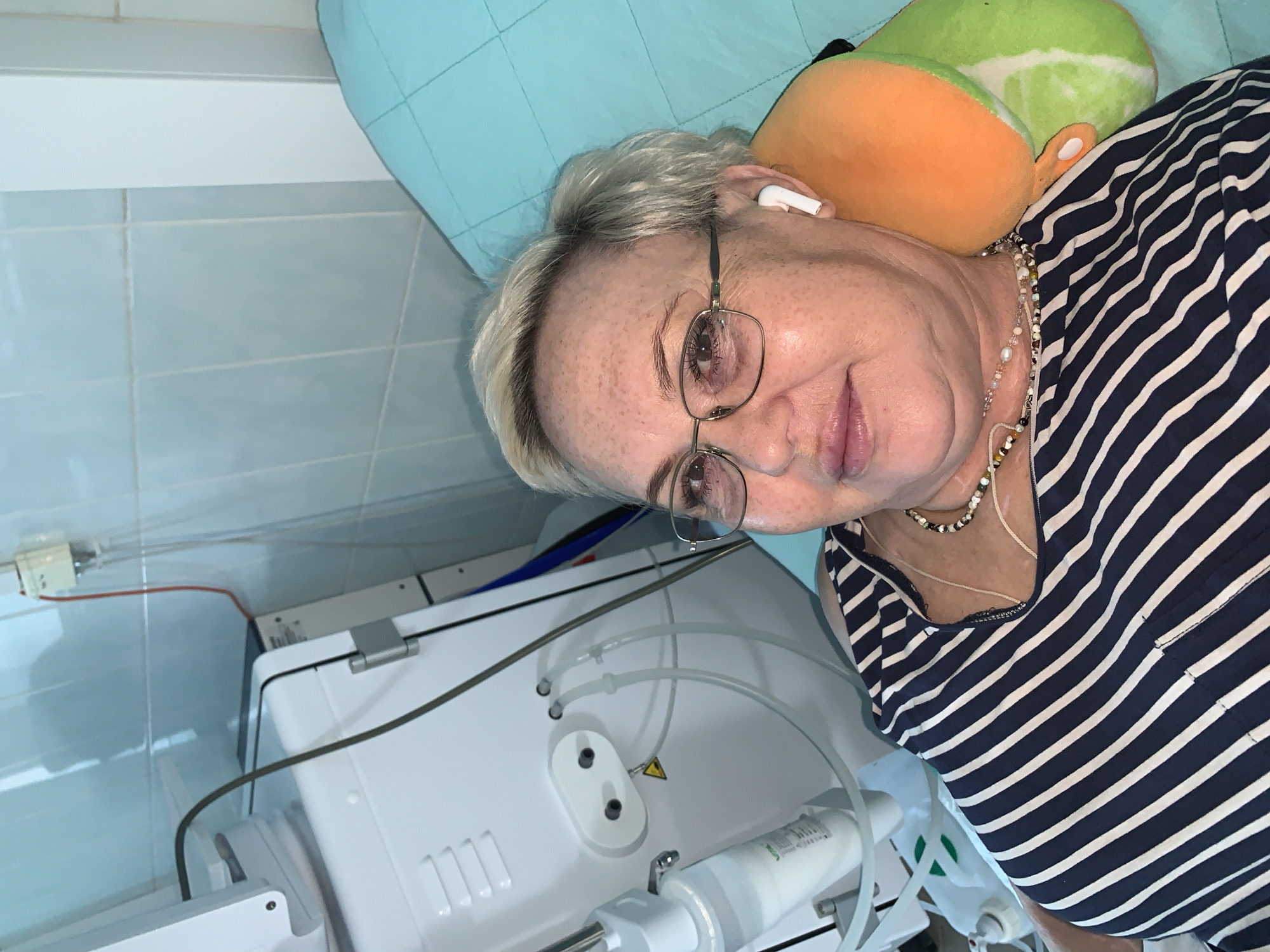 A medium shot depicts an older woman with short, light-colored hair and glasses, lying on her side with a stuffed animal behind her head. She wears a navy and white striped shirt and a necklace. An earbud is visible in her left ear.
Next to her is a white medical device with tubes and a power outlet, suggesting a medical setting. The background features blue-green tiled walls. The location is unspecified but given the prompt it is in the city of Chaikovsky, Russia.