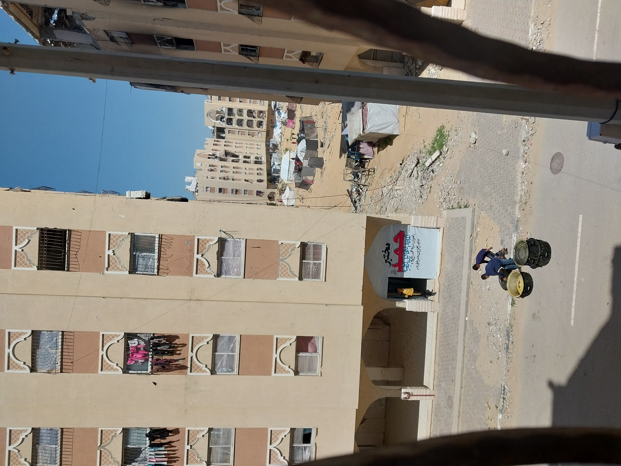An eye-level, outdoor shot shows a beige apartment building on the left, with a view of a camp of tents, another taller apartment building in the background, and a street on the right. Two men are walking down the street, carrying a large object.
The apartment building on the left has several floors, each with multiple windows. Laundry hangs from one of the windows. A banner with Arabic writing hangs on the side of the building.
The camp in the background appears to be a collection of temporary shelters and tents. The taller apartment building in the background is mostly tan.
The two men are wearing dark clothes and walking along the right side of the street, carrying what appears to be a large, black and yellow cylindrical object.
The sky is clear and blue. The photo is taken from a high vantage point, likely from another apartment building.