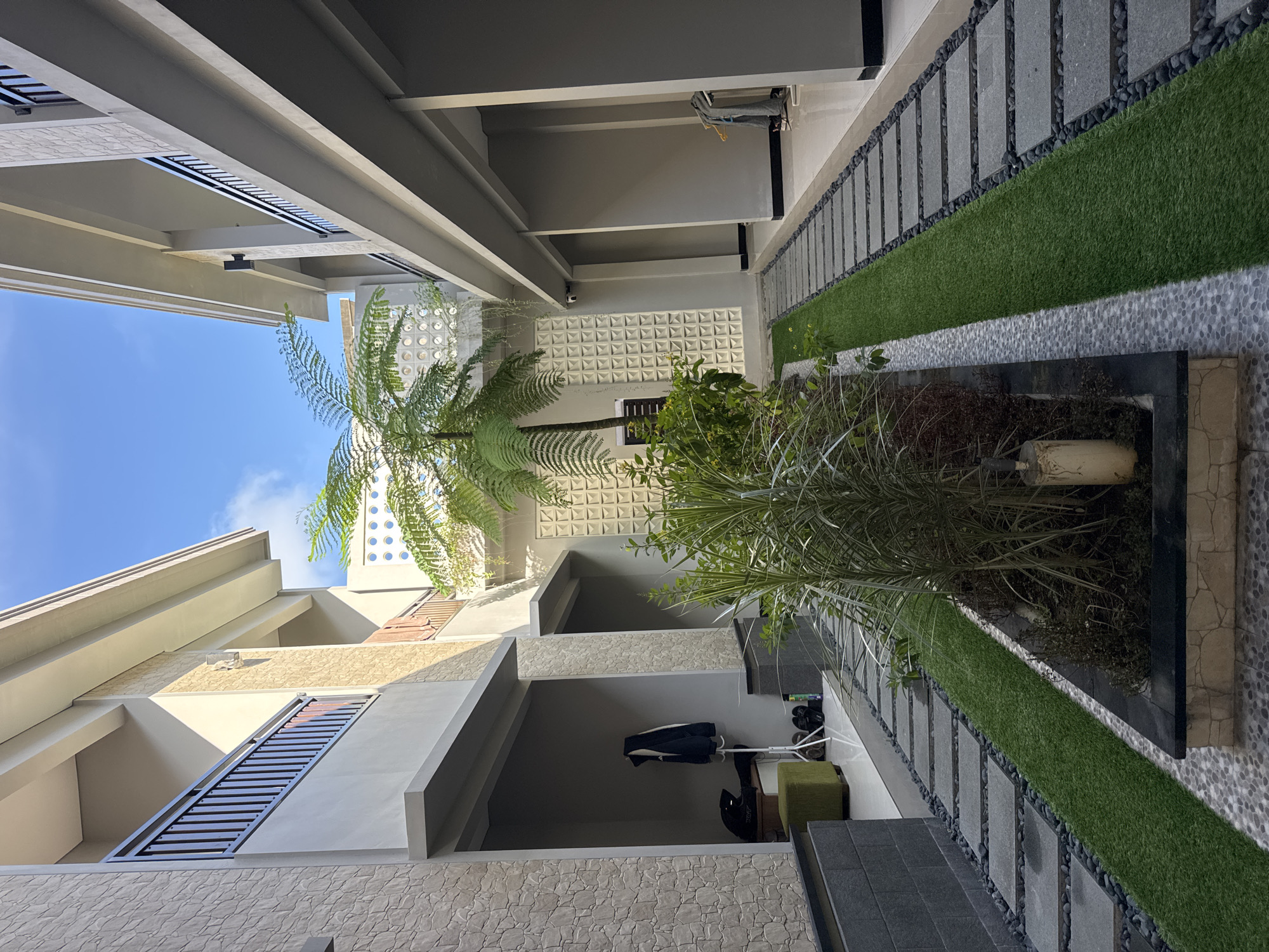 The image shows an outdoor courtyard in Kuta, Indonesia, featuring modern architecture and landscaping. Two trees, one a tree fern and the other a type of palm, are prominent in a raised planter bed. A pathway runs alongside the planter, constructed with alternating gray stone slabs and artificial turf, bordered by small black pebbles. The buildings surrounding the courtyard have a textured stone exterior and concrete beams. A blue sky is visible overhead. A small storage area beneath one of the upper floors contains some items. A patterned concrete wall is visible in the background.