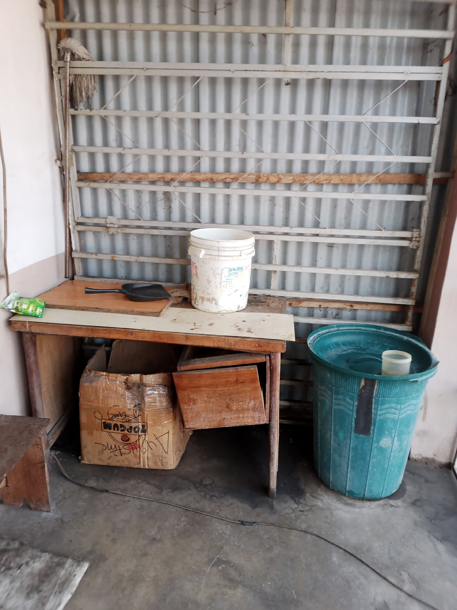 An exterior view captures a cluttered outdoor area, possibly a yard or alley, in Jalingo, Nigeria. A wooden table, stained and weathered, occupies the center left, with various items scattered across its surface and underneath. A black dustpan rests on the table, next to a dirty white plastic bucket. Underneath the table, cardboard boxes and wooden crates are piled haphazardly. To the right of the table stands a large, teal plastic trash can. The background features a corrugated metal wall with a white metal rack structure in front of it. A mop with a white head hangs from the rack on the far left. The ground is concrete, with some damp patches and a dark electrical cord snaking across it. The scene appears unkempt and utilitarian, suggesting a storage or work area.