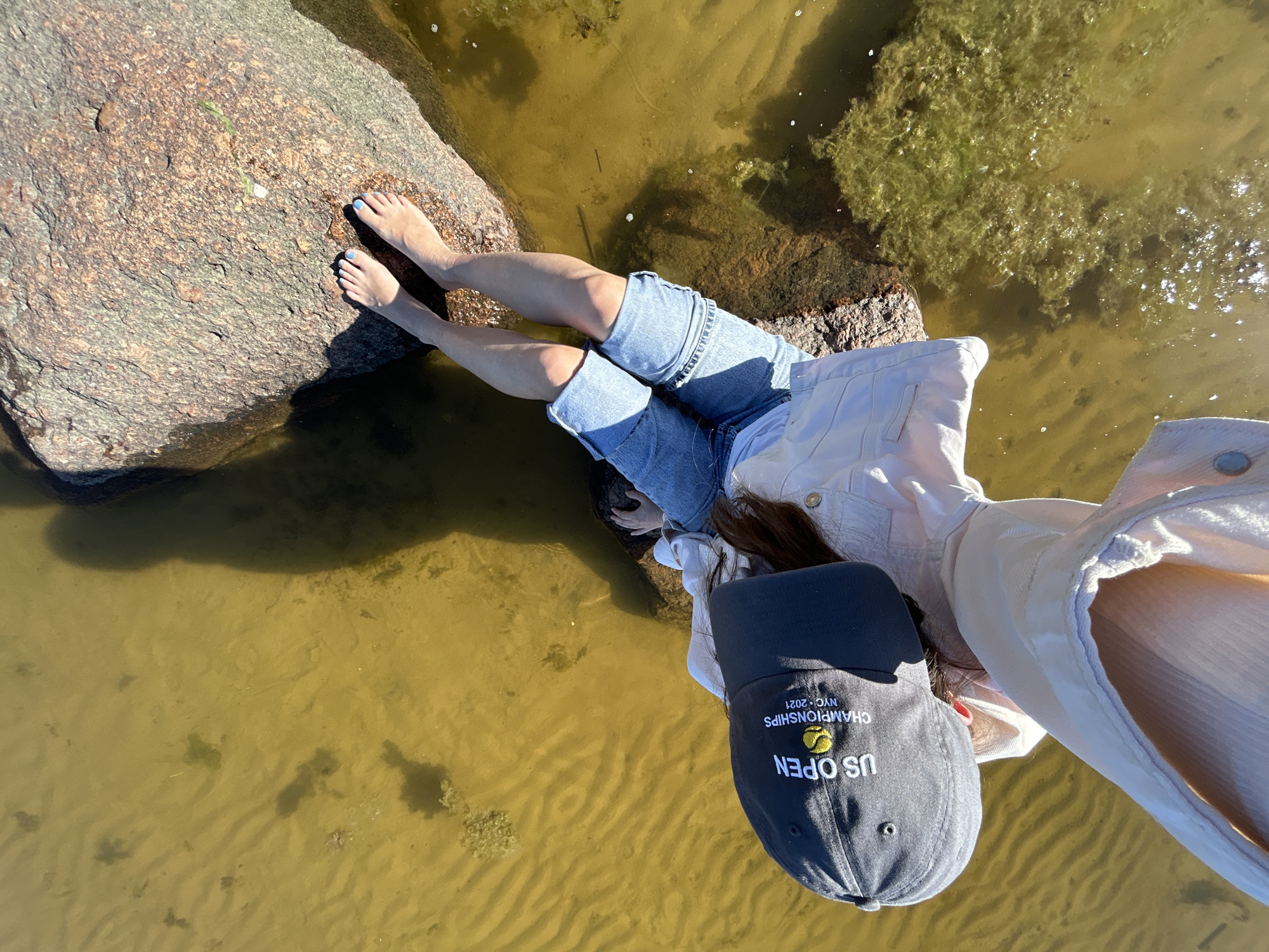 An overhead perspective shot shows a person seated on a large rock in murky, shallow water with visible algae. The individual, wearing a US Open baseball cap, a light-colored jacket, and denim shorts, has their legs extended into the water. Their feet, with blue-painted toenails, rest on the rock. The water appears calm with ripples and shadows indicating sunlight. The scene is set outdoors, with the rock and water suggesting a natural environment. The city of Shepelyovo, Russia, is mentioned as the location, though no specific landmarks confirming this are visible in the image.