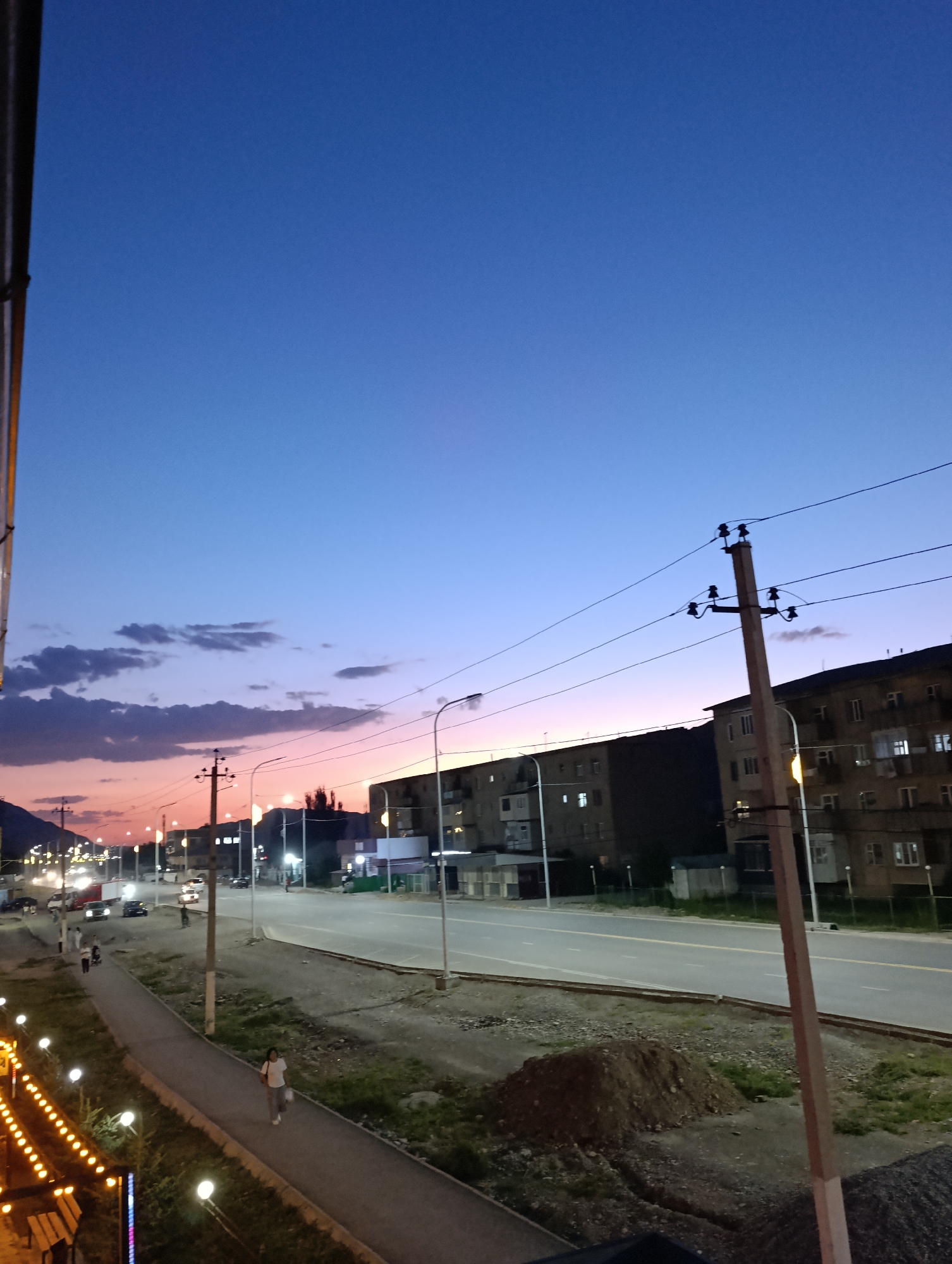 The image depicts a twilight scene in the city of Naryn, Kyrgyzstan. The sky transitions from a deep blue at the top to hues of pink and orange near the horizon, indicating either dawn or dusk. A wide paved road runs through the scene, with cars and streetlights visible. To the right, a multi-story building with illuminated windows stands. On the left side of the road, a pedestrian walkway is bordered by decorative lights and benches. Several people are walking on the walkway. Power lines and utility poles are prominent throughout the frame, with one particularly large pole in the foreground. In the distance, mountains can be seen on the left side of the image. The overall impression is that of an urban environment settling into the evening.