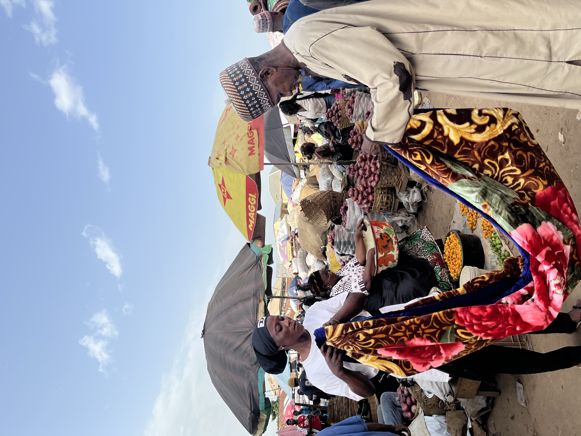 An outdoor market scene in Rafin Par, Nigeria, features people engaged in commerce. In the foreground, a woman in a white t-shirt and black headscarf holds up a colorful blanket with a floral and ornate pattern. To her right, a man in a light-colored robe and a patterned cap examines a similar blanket. Behind them, various stalls are visible, some with large umbrellas providing shade. Baskets of red onions and other produce are displayed. The sky above is partly cloudy.