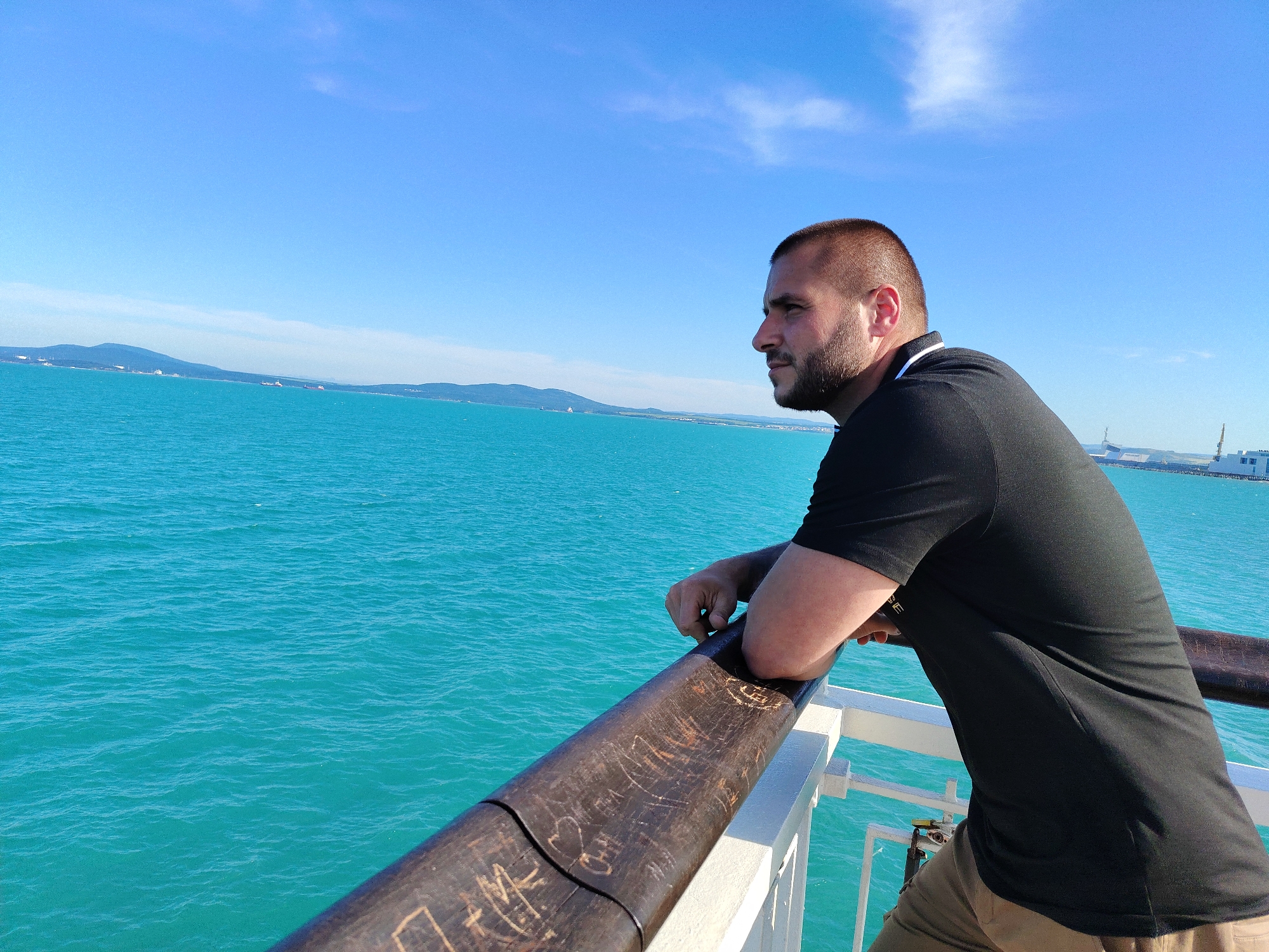 A medium shot shows a man on a boat, leaning on a wooden railing and looking out at the turquoise water. The man has short dark hair and a beard, and is wearing a black t-shirt and tan pants. The sea stretches out to the horizon, where a hazy landmass with hills and buildings is visible. The sky is a clear blue with a few wispy clouds. The wooden railing has carvings on it, suggesting it's a well-used part of the boat. The overall scene suggests a peaceful moment at sea, possibly near the city of Burgas, Bulgaria, given the distinctive color of the water and the coastal landscape.