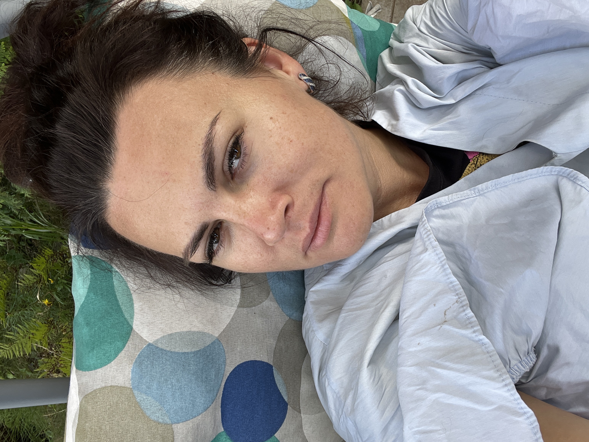 A woman is lying on her back on a pillow with a circular pattern, wrapped in a light-colored garment. She is looking towards the camera with a neutral expression. Her hair is pulled up into a messy bun. In the background, greenery is visible, suggesting an outdoor setting, possibly in Роса Снт, Russia. The lighting indicates it is daytime.