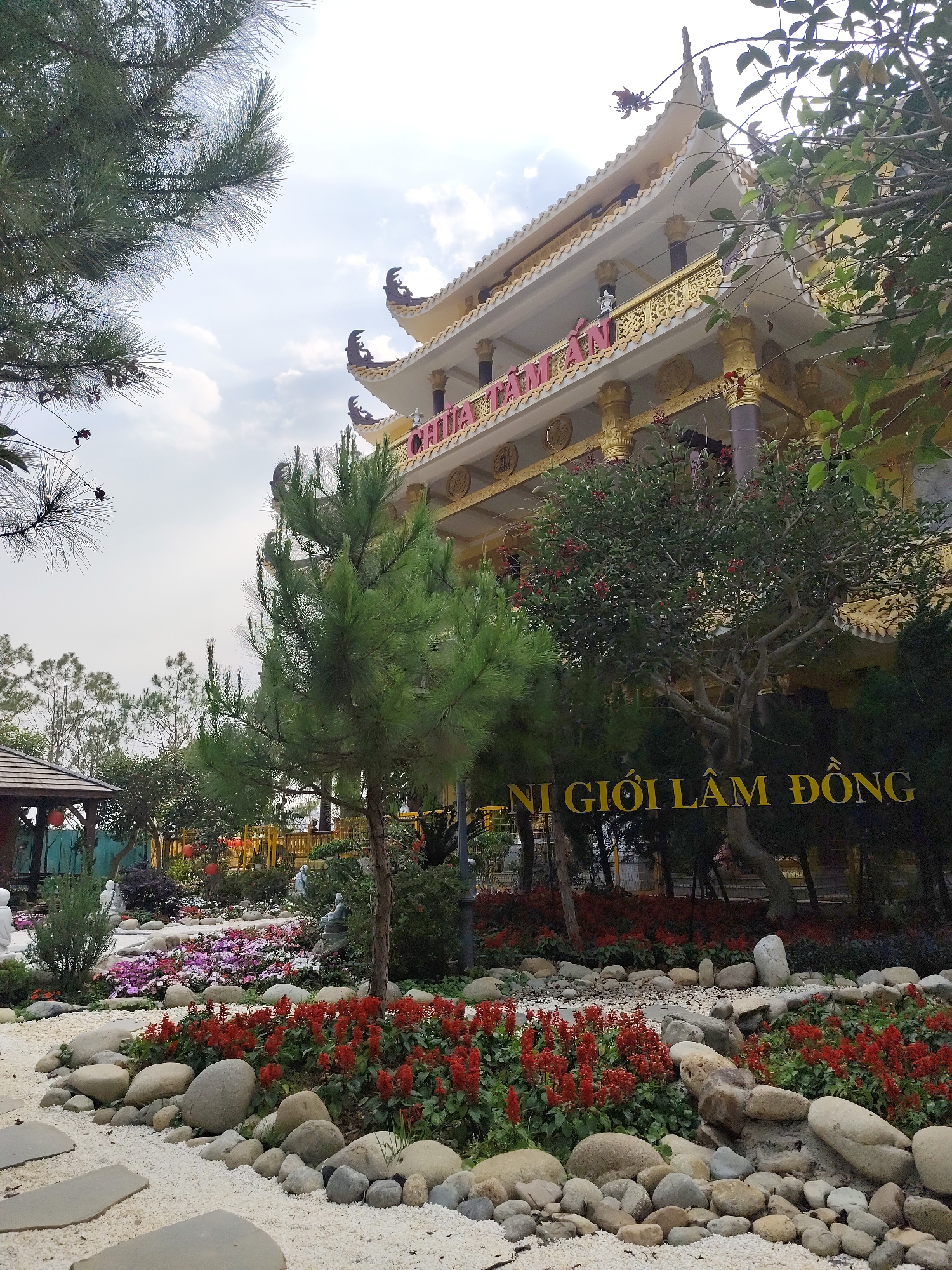 The image shows a building within the grounds of the Truc Lam Pagoda complex in Da Lat, Vietnam. The pagoda building is multi-tiered with a cream-colored exterior and gold trim, topped with decorative figures. Red lettering, partially obscured, reads "CHÙA TÂM AN." A large, yellow sign is partly visible, reading "NI GIỚI LÂM ĐỒNG."
The foreground features a landscaped garden with various plants, red flowers, and stones lining a white gravel path. A small gazebo structure is visible in the background on the left. Trees and foliage surround the pagoda and garden. The sky is overcast with light clouds.