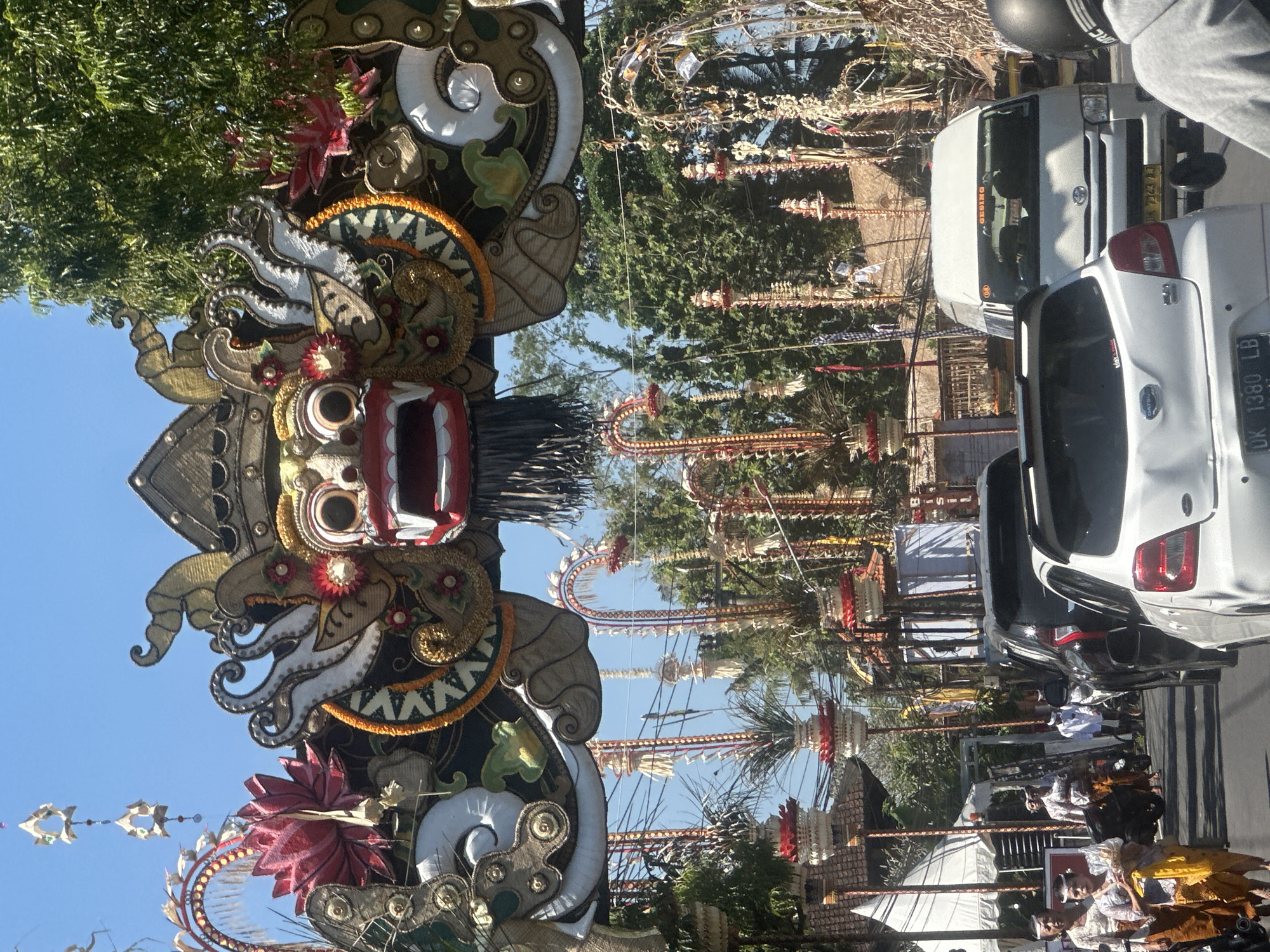 The image shows a street scene in Sukawati, Indonesia, on a clear, sunny day. An elaborate, decorative archway, featuring a large, ornate mask with red and white teeth and wide eyes, spans the road. The archway is adorned with gold, black, and white embellishments, as well as red and pink lotus-like decorations.
Below the archway, various festive decorations line the street, including tall, slender poles topped with tiered, red and white structures and woven elements. Hanging from some of these poles are small, decorative items. Trees with lush green foliage form a backdrop behind the decorations and archway.
The street itself is paved and appears to be a busy thoroughfare. Several vehicles are present, including a white Toyota van with the license plate "DK 7943 LB" and the word "GESING" visible on the windshield, and a white hatchback car with the license plate "DK 1380 LB". A black SUV is also visible.
On the left side of the road, near the foreground, several people are walking. Two women are dressed in traditional Balinese attire, consisting of patterned tops and sarongs or sashes in brown and yellow. One woman wears a white headband. Another person in the background, also in traditional clothing, appears to be walking away from the camera. A motorcyclist wearing a helmet is in the far right foreground, facing away from the camera. Tents or canopies are set up on the left side of the street, suggesting a possible market or event. The overall atmosphere appears to be festive and possibly related to a local celebration or ceremony.