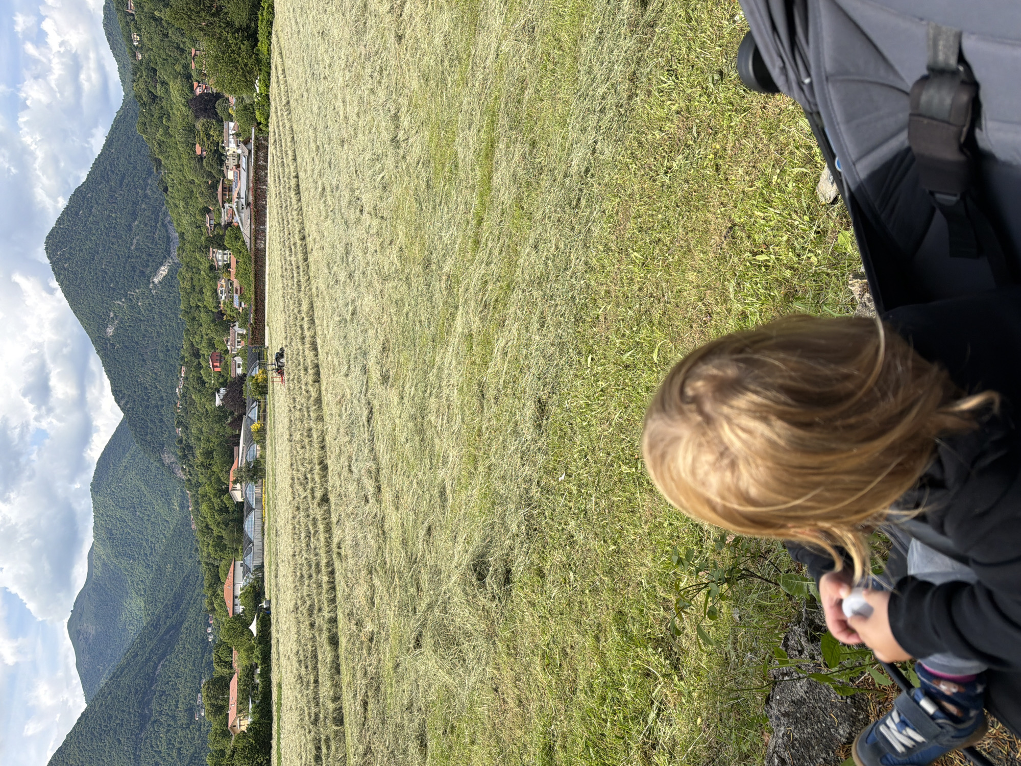 A child with blonde hair is shown in the foreground. The child is partially obscured by a dark-colored object, possibly a stroller. The child is holding something white in their hands. The child is wearing blue shoes. Behind the child, there is a field of cut grass, partially green and partially dry. In the background, there are mountains and a row of buildings. The sky is partly cloudy. The location may be Penzano, Italy.
