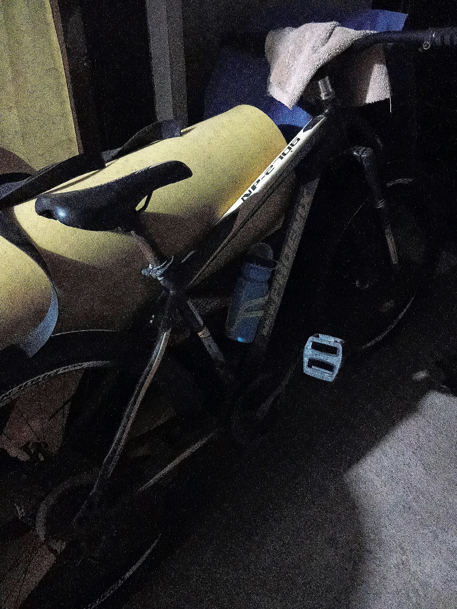 The image depicts a bicycle in what appears to be a storage area. The bicycle is primarily dark-colored, with a yellow-cushioned seat and a white frame with some text that is partially visible and difficult to decipher. A blue water bottle is attached to the frame. The background is dark and out of focus, suggesting a dimly lit environment. Part of a grey, textured surface, possibly concrete or asphalt, is visible in the lower right of the frame. There is a folded cloth or blanket draped over the handlebars. The scene is static, with no people or ongoing activity visible. There is no information to determine the location within the city of Cikal, Indonesia, as the image only provides a close-up view of the bicycle and its immediate surroundings.