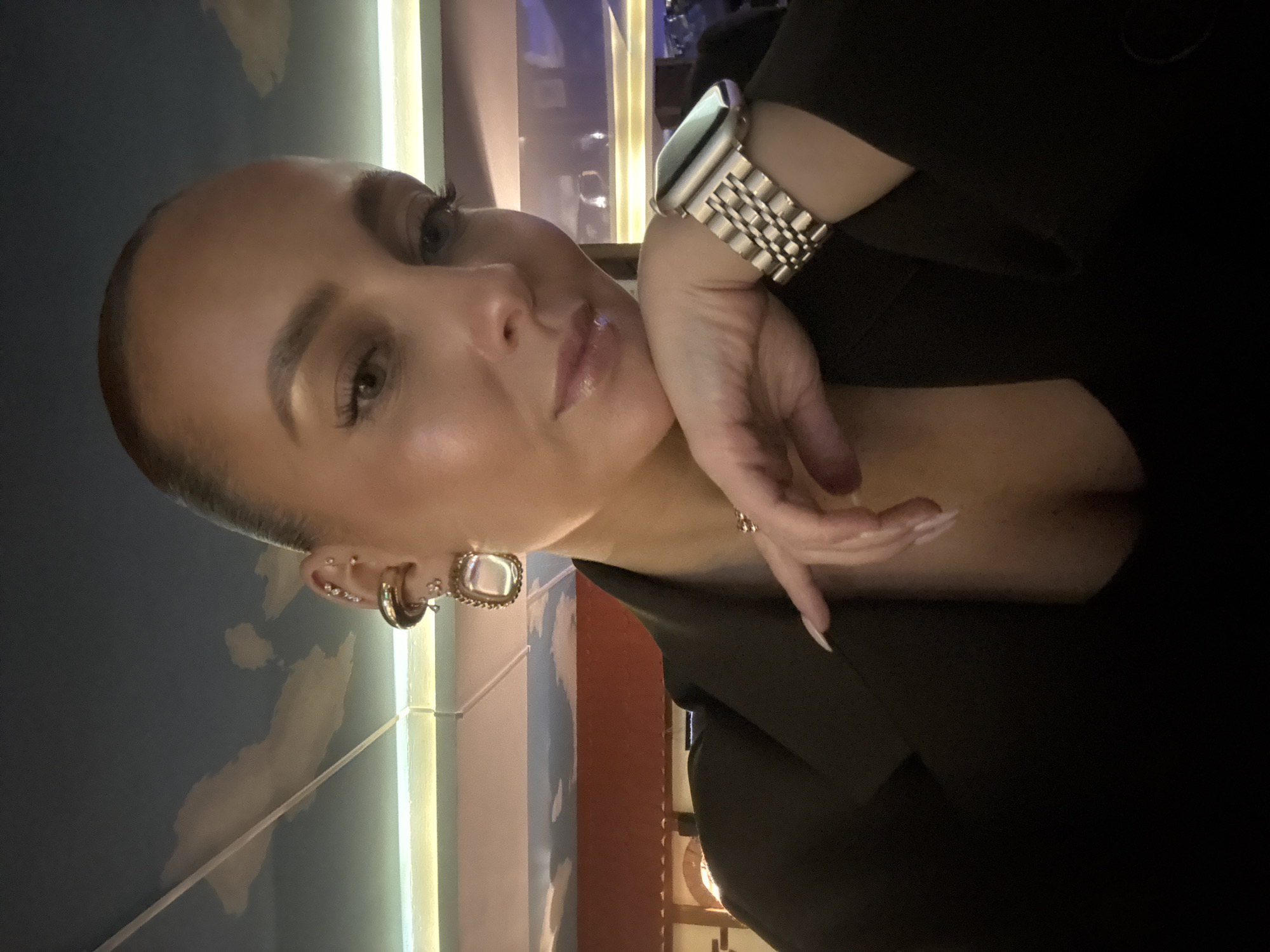 A female individual is depicted in a close-up, chest-up portrait within an indoor establishment in Chaikovsky, Russia. She has light skin, visible makeup, defined eyebrows, and extended eyelashes. Her lips are glossy, and she has a labret piercing on her lower lip. Her hair is styled back, and she is looking directly at the camera with a slight smile. She is wearing a dark blazer over a dark top, revealing a moderate amount of cleavage. Her left ear is adorned with multiple piercings, including a large gold-toned hoop with an ornate square-shaped dangling earring on the lobe, and several smaller studs and hoops on the cartilage. A gold-toned metal link watch is visible on her left wrist. Her left hand is positioned with her chin resting on her knuckles, and her fingers, adorned with long, pointed nails with light polish, are slightly curled. The background is dimly lit and out of focus. A painted ceiling depicting a blue sky with white clouds is visible at the top. Below this, a horizontal light strip emits a greenish-yellow glow, situated above a reddish-brown architectural element, possibly a beam or wall section. Distant blurred lights and reflections suggest an indoor public space, likely a restaurant or bar.